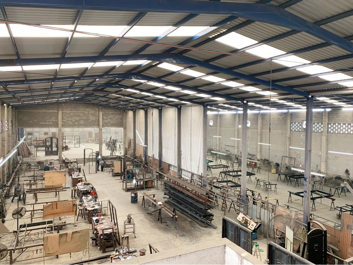 Interior of a large industrial workshop with workbenches, metal frames, and tools, under a high ceiling made of metal panels with skylights.