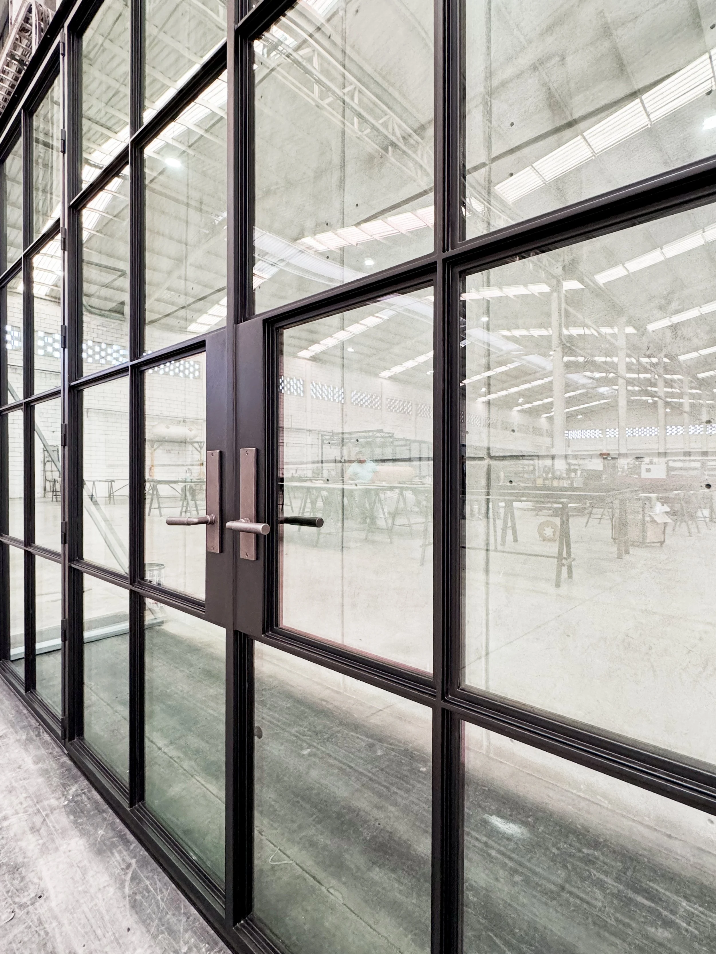 A glass door with black metal framing, handles, and a latch, leading into an industrial or commercial space with a high ceiling and exposed structural elements.