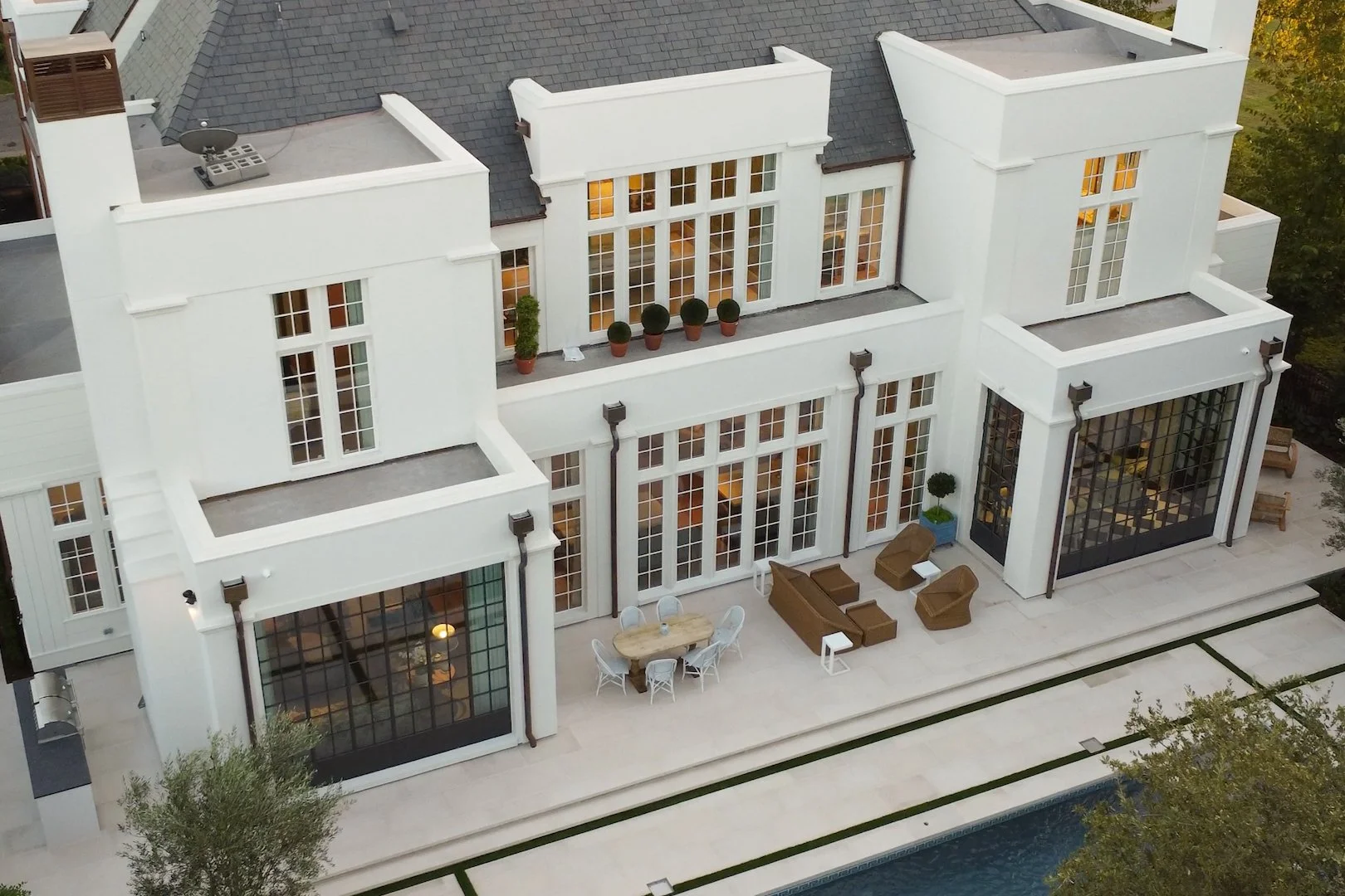 Aerial view of a modern white multi-story house with large glass windows, outdoor patio furniture, potted plants, and a swimming pool.