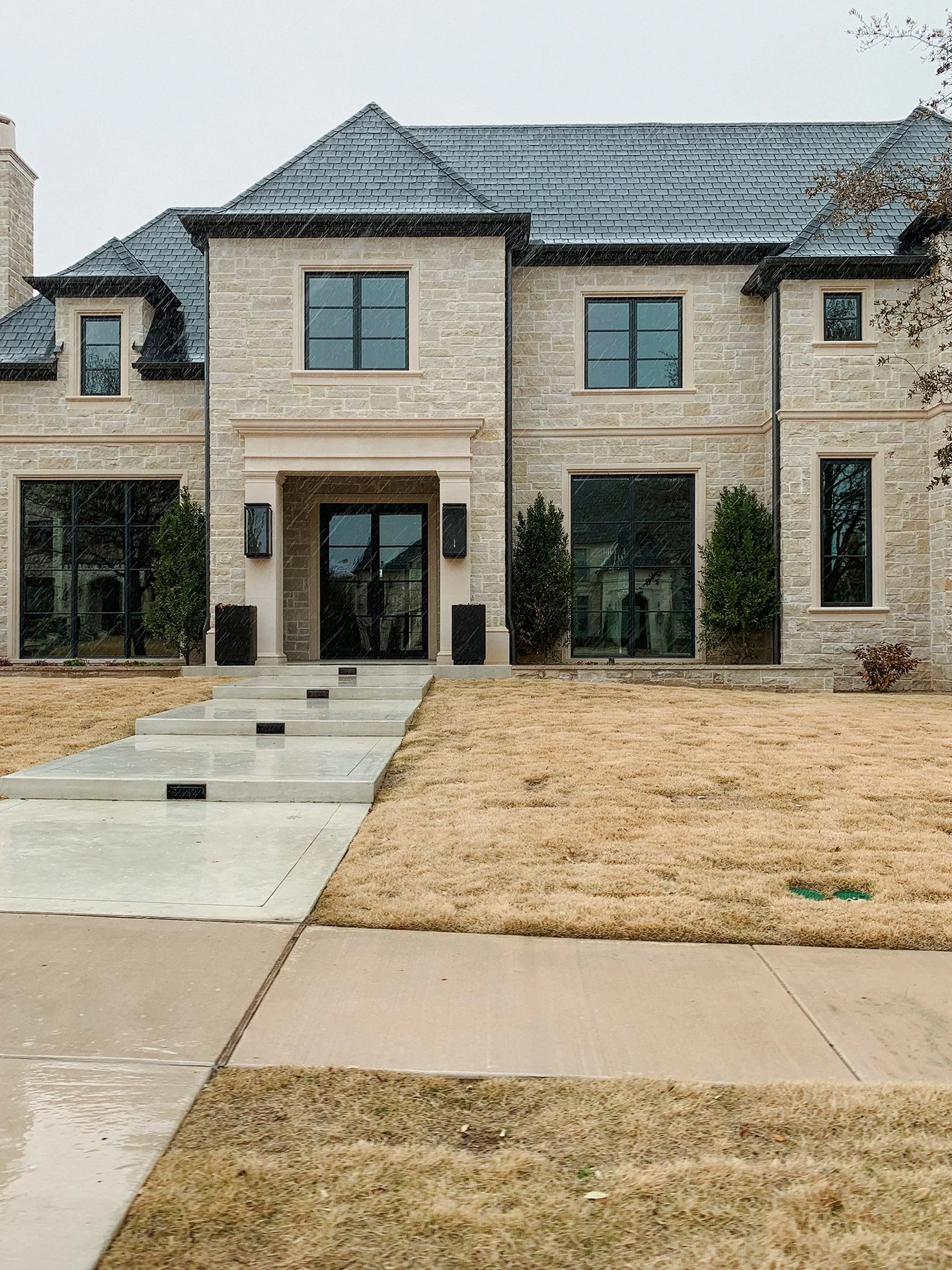 Front view of a modern residential house with beige stone exterior, black window frames, and a blue tiled roof. A concrete sidewalk runs in front, leading to the porch with stairs. The lawn is dry and brown, and there are small bushes near the house.