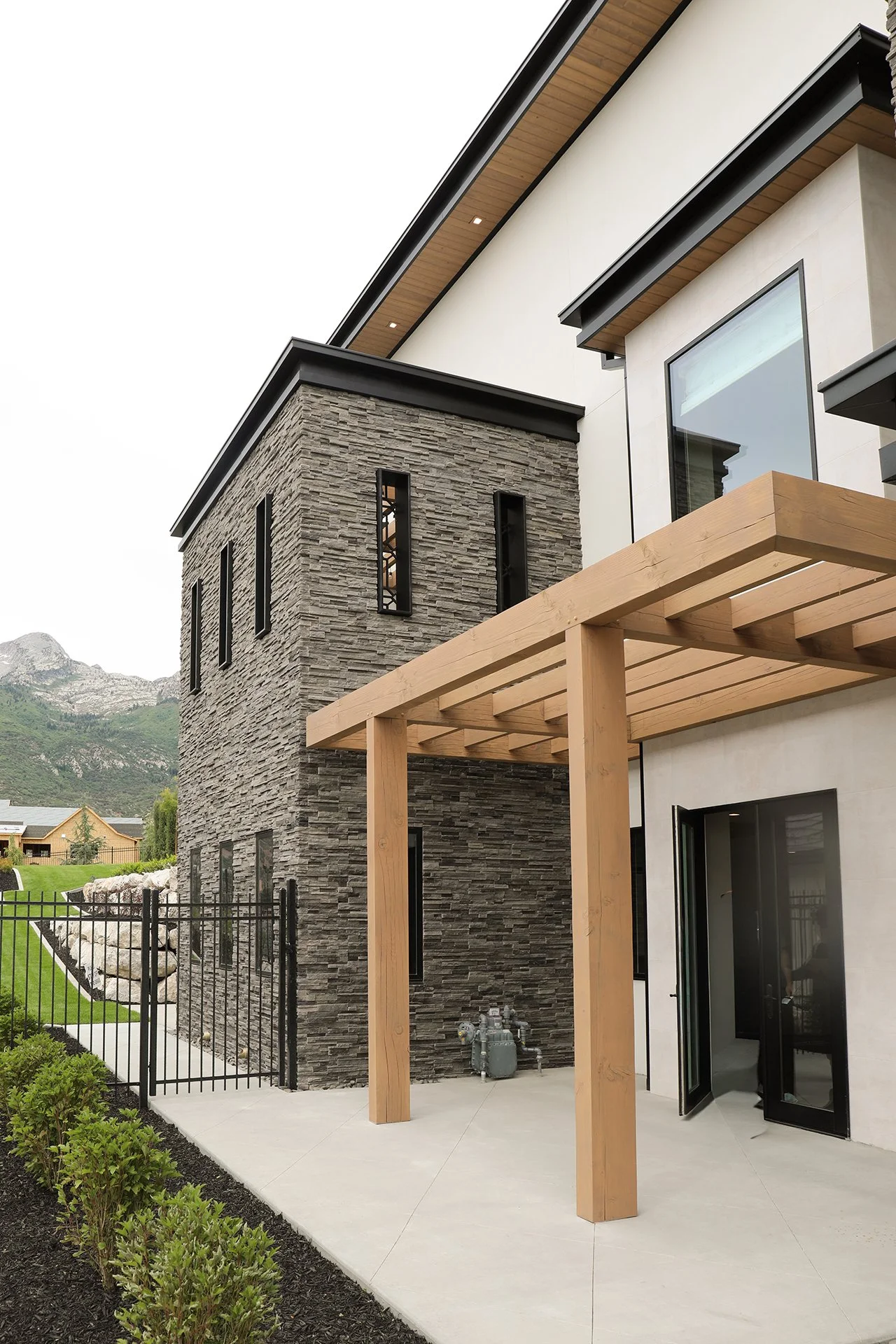 Modern house with a stone tower, black metal windows, wooden pergola, and sliding glass doors, set against a mountainous background.