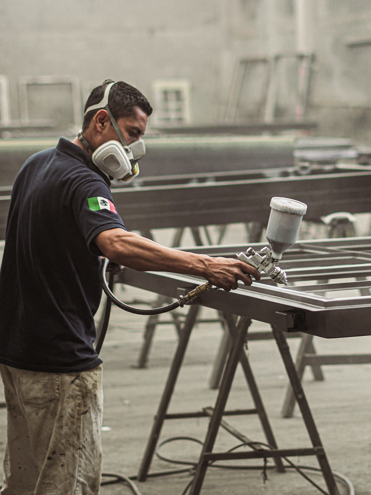 A man wearing a black shirt with a Mexican flag patch on the sleeve is spray painting a metal surface in an industrial workshop. He is using a spray gun and wearing a respirator mask for protection.