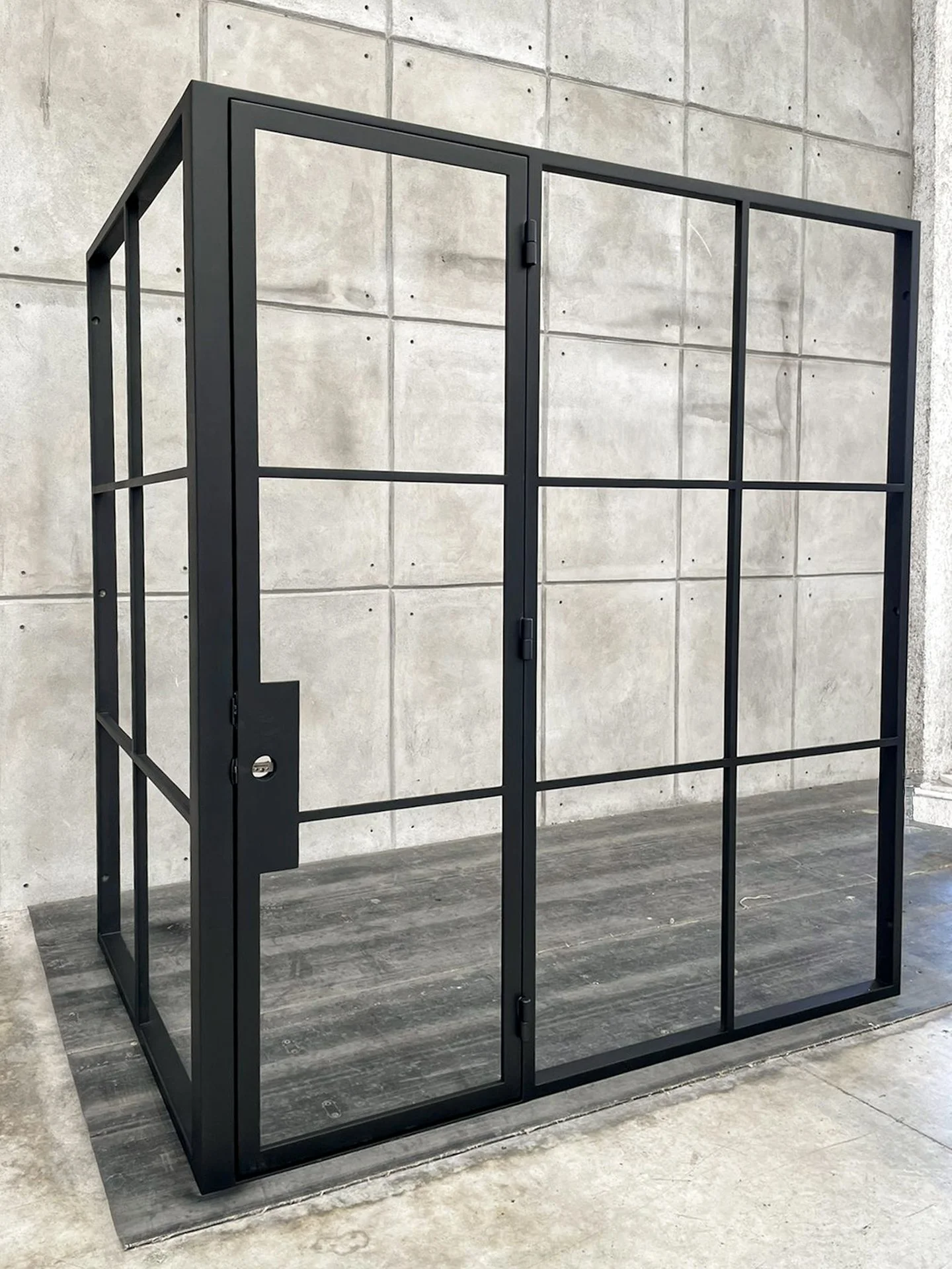 A black metal frame with glass panels, presumably a door or partition, standing indoors against a concrete wall.