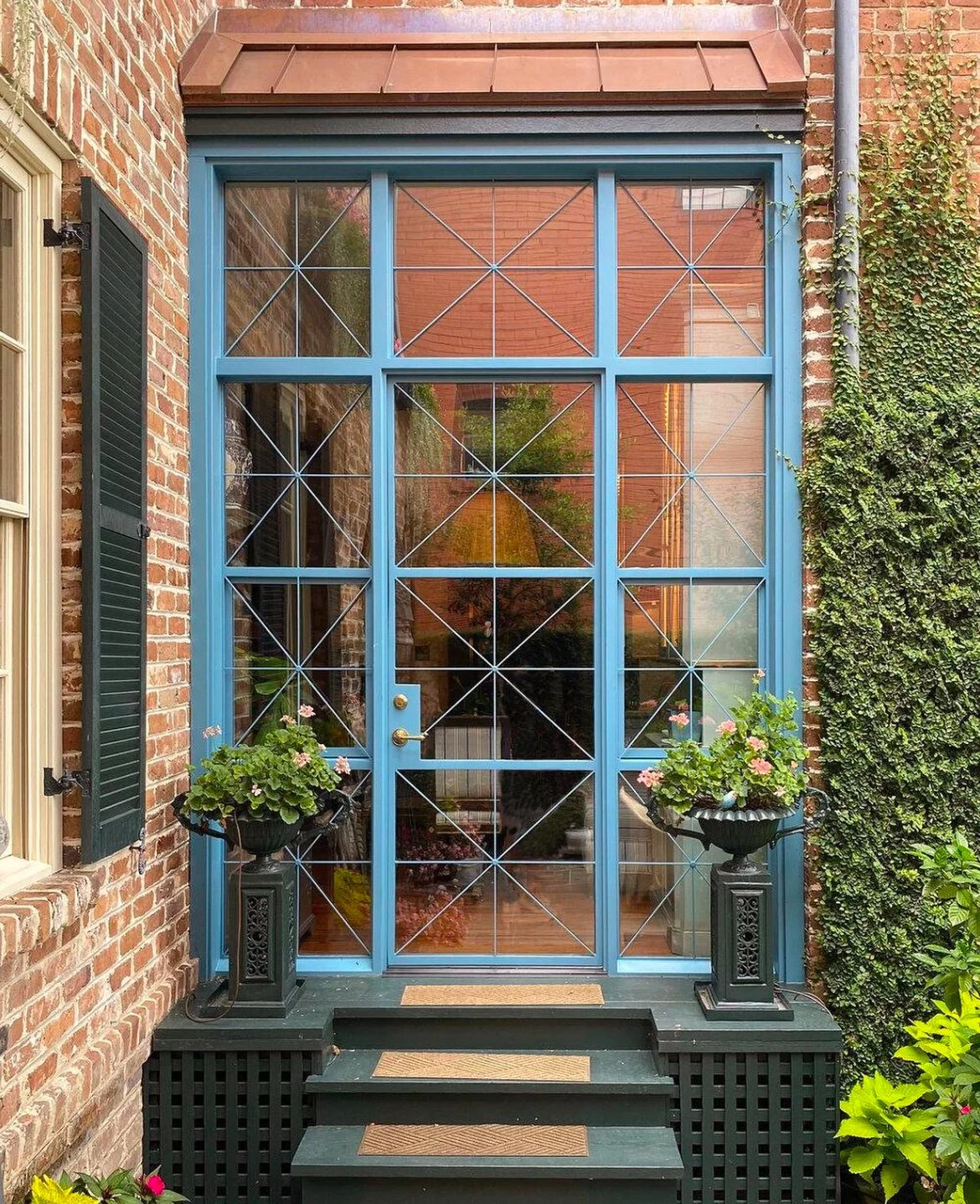 Front porch with a glass and blue metal door, pink flowers in black planters on black pedestals, brick walls, green ivy on the right side.