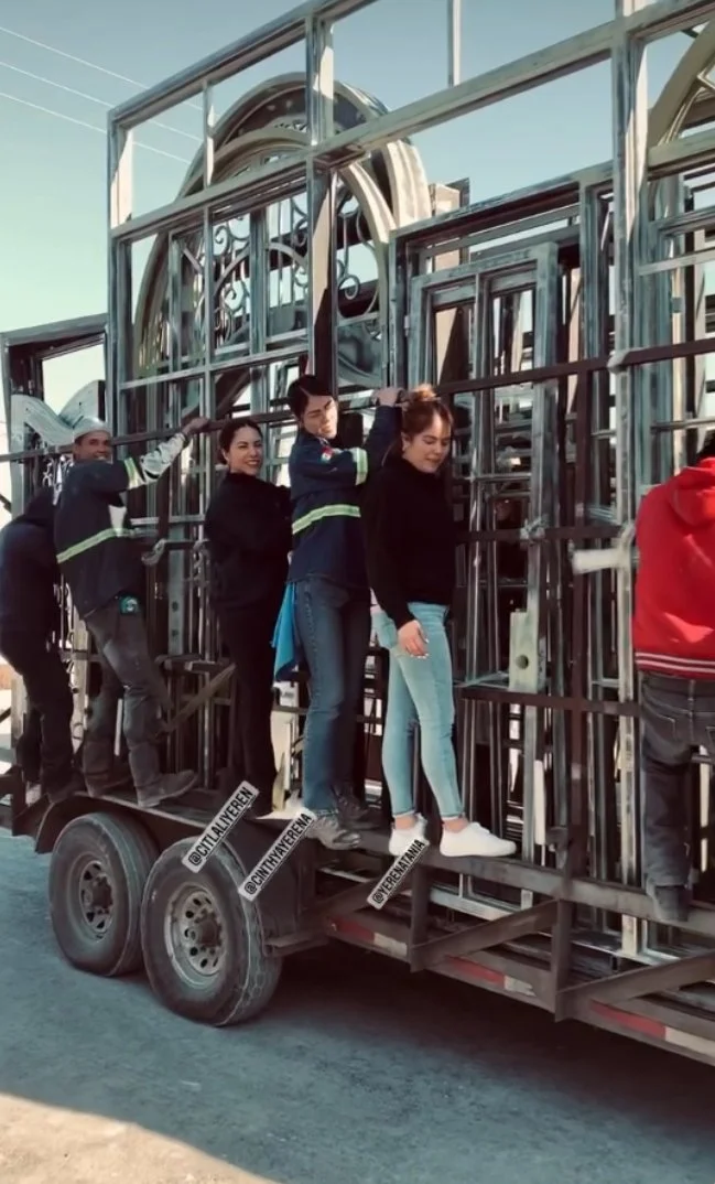 People standing on a trailer loaded with metal structures, with a large decorative wheel in the background.