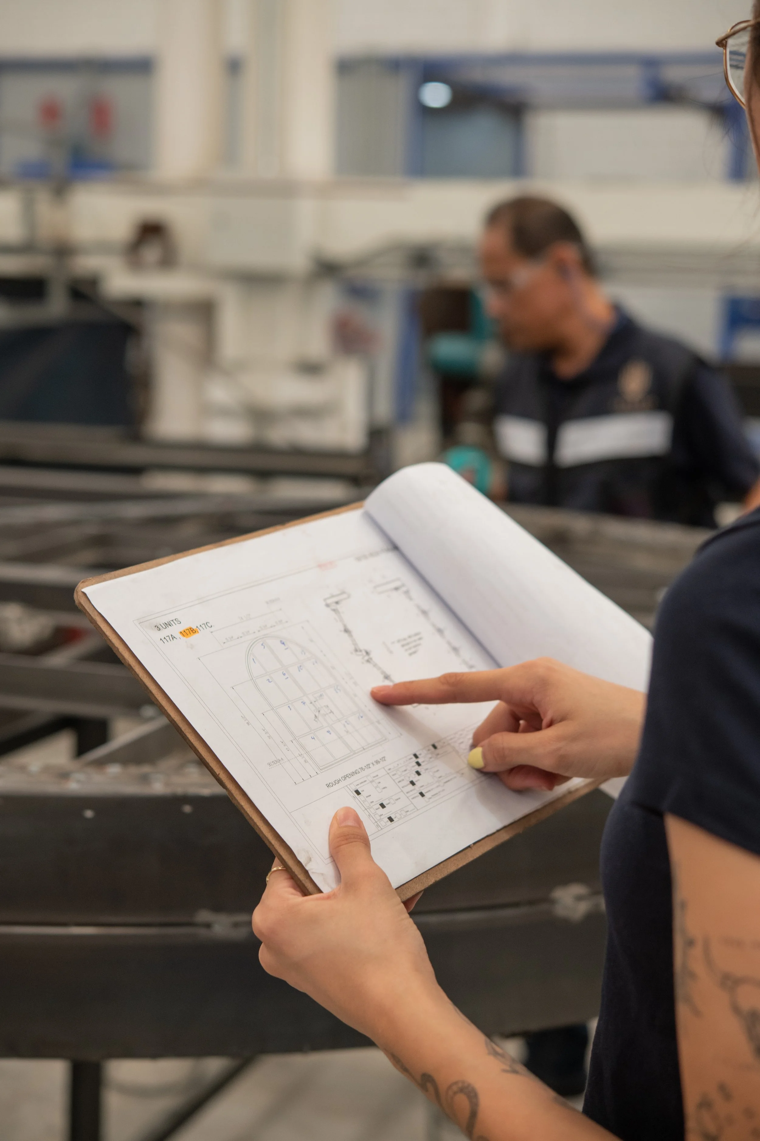 Person holding a clipboard with technical drawings or blueprints, focusing on the paperwork, with an industrial setting in the background and a blurred person working.