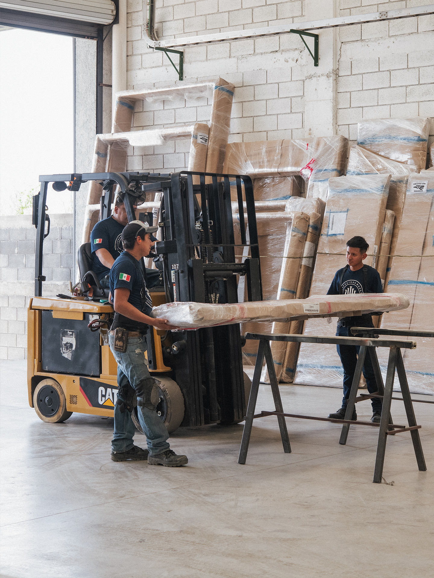 Three workers are moving large wrapped items in a warehouse. Two are operating a forklift, while one stands nearby.