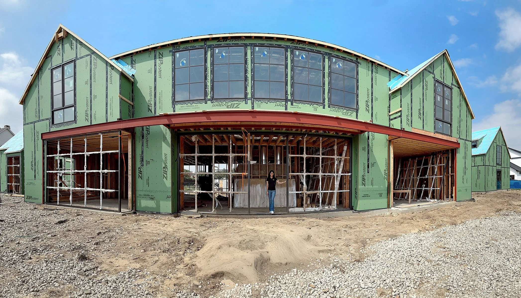 Under construction house with green sheathing, a large front opening, and worker standing inside, with a blue sky above.