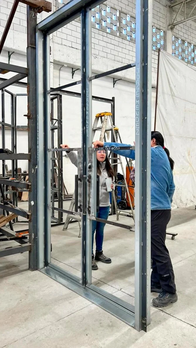 Two women are working on a metal door frame in a large industrial workshop. One woman holds a cup, the other points at the frame while standing near a ladder. The background contains scaffolding, a white wall, and equipment.