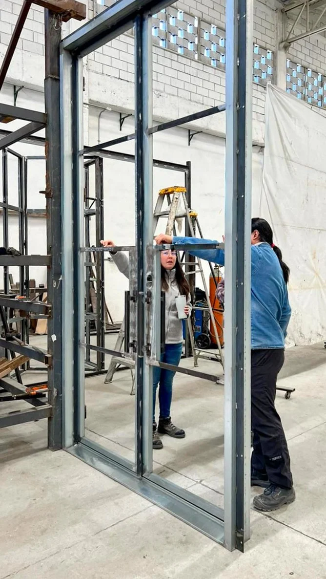 Two women are inside a steel frame structure in a workshop, with ladders and tools visible around them.