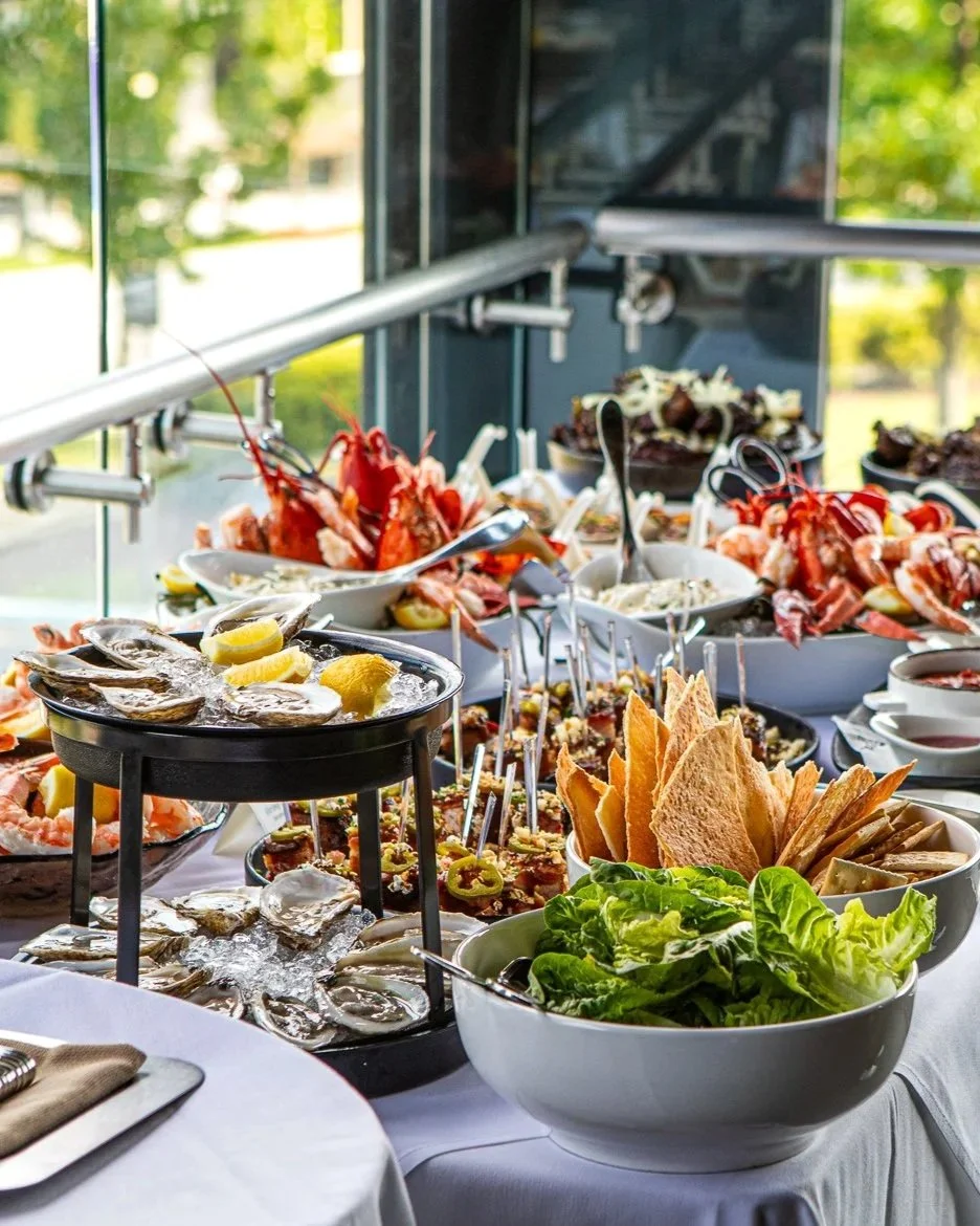 A seafood buffet with oysters on ice, lobster, shrimp, crackers, lettuce, and various dipping sauces, set on a table near large windows.