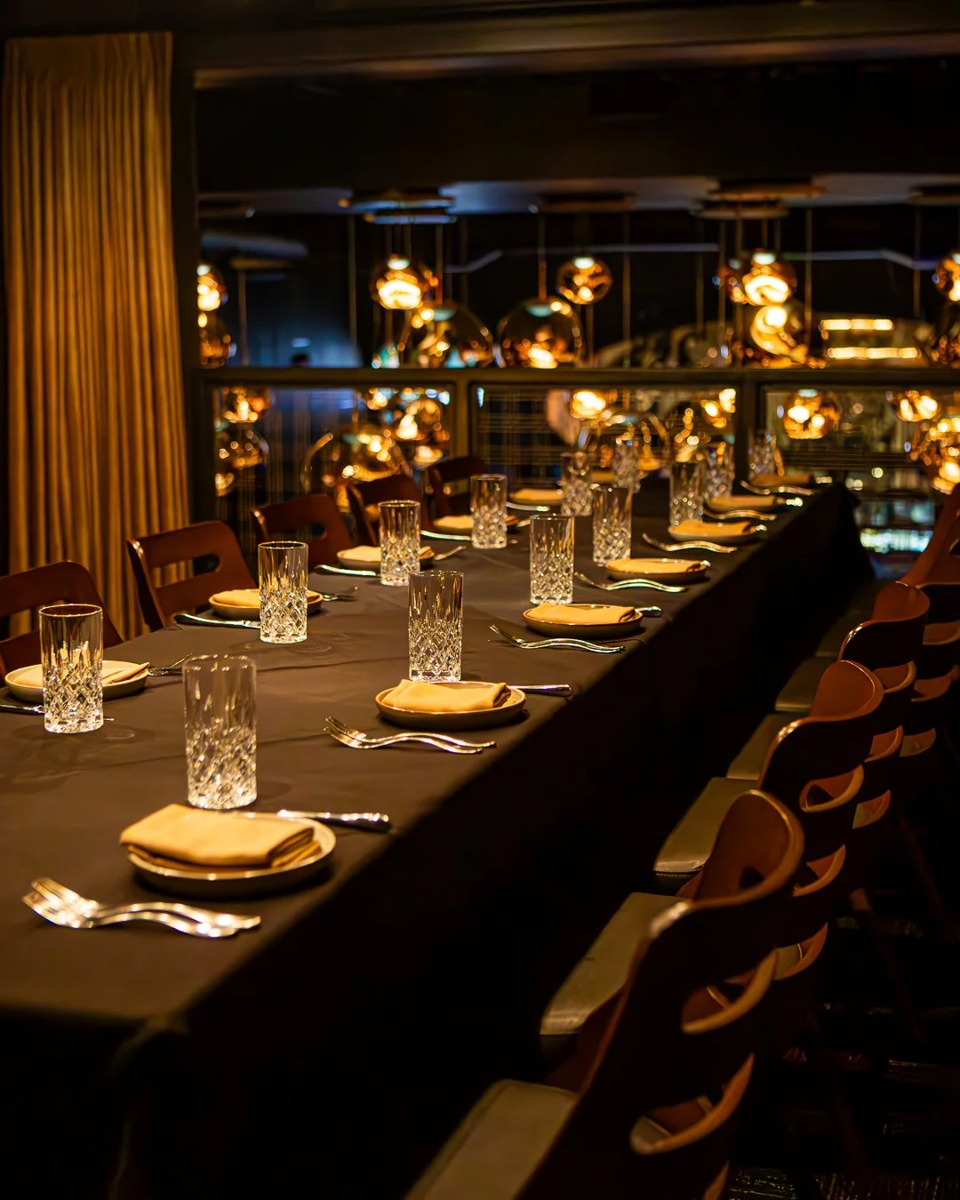 Elegant dining table set for a formal dinner with glasses, plates, silverware, and napkins, illuminated by warm hanging pendant lights in a dimly lit restaurant or banquet hall.