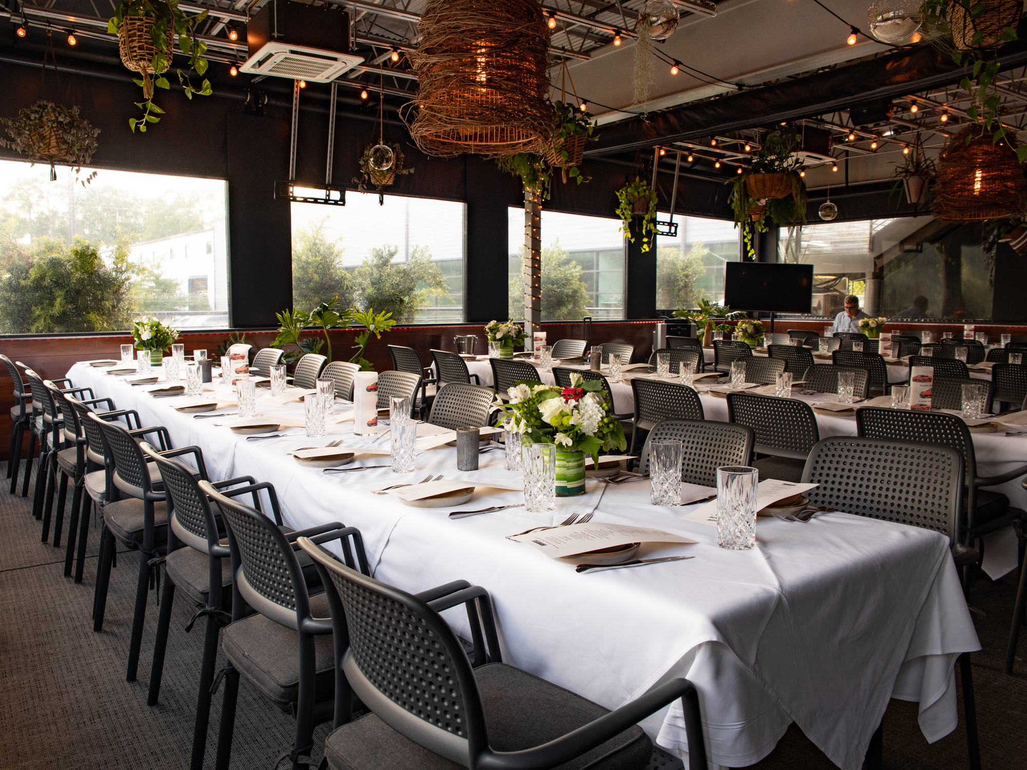 Long dining table with white tablecloth, set with glasses, plates, napkins, and floral centerpieces in a decorated restaurant or event space with natural light, hanging plants, and ambient lighting.