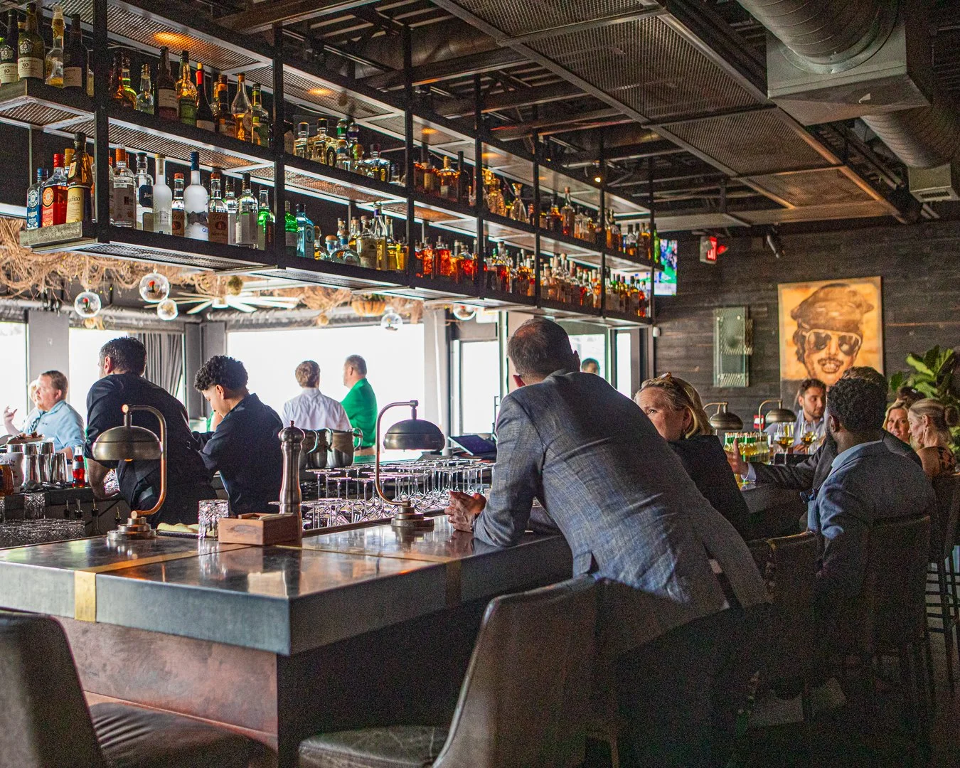 Interior of a modern bar with people sitting at the bar counter, shelves stocked with bottles of liquor, and a large window letting in natural light. There is artwork on the wall, and the atmosphere appears lively and stylish.