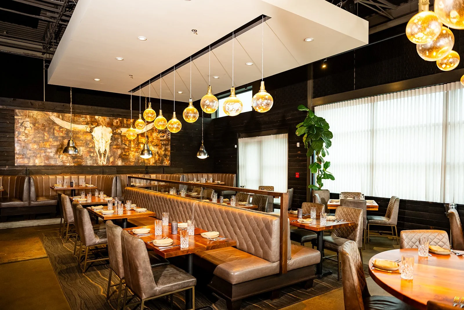 Interior of a modern restaurant with tufted leather booths, wooden tables, and hanging pendant lights, featuring a decorative animal skull painting on dark wooden walls and a large potted plant by windows with white blinds.