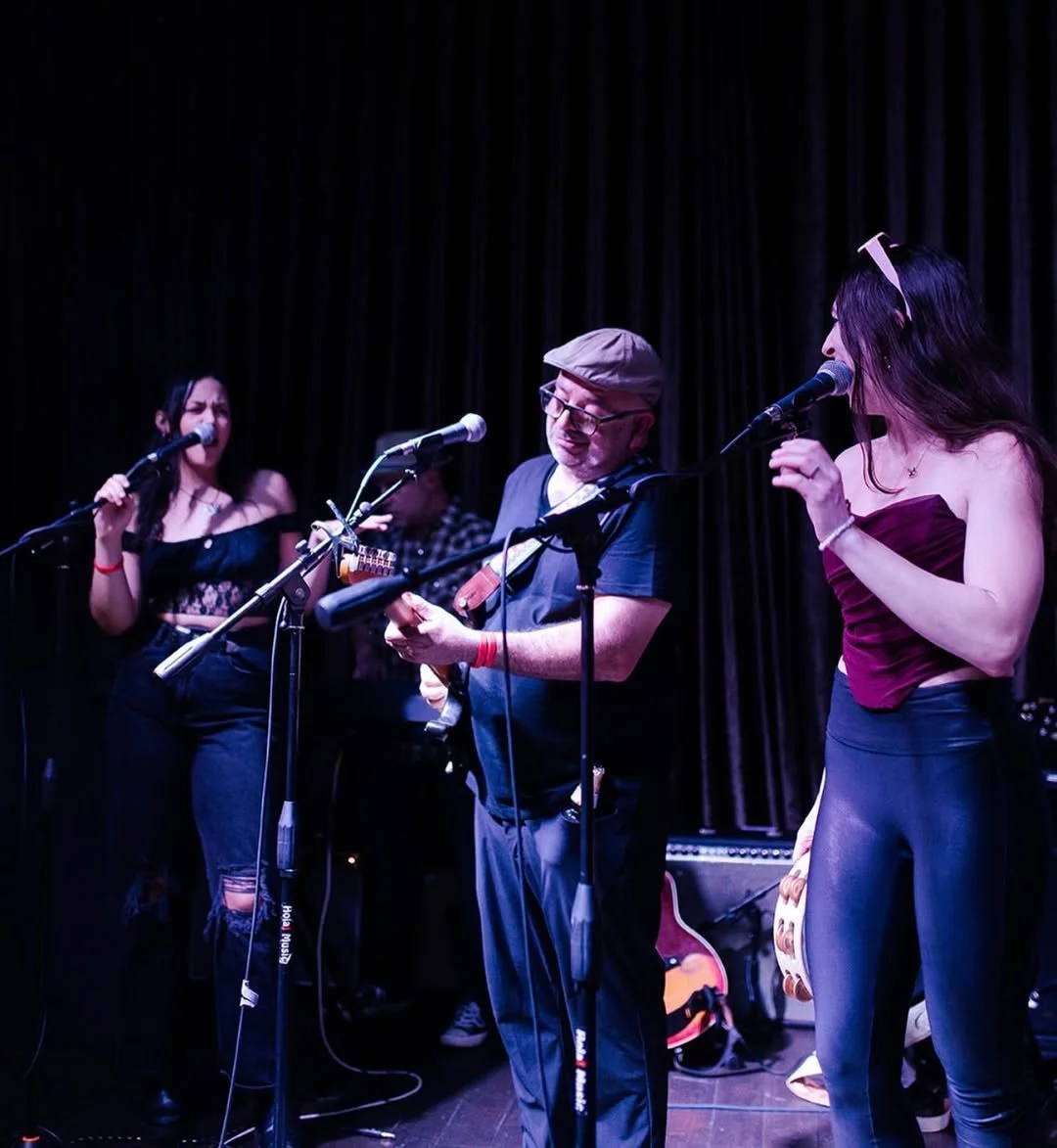 A band performing on stage with three women singing into microphones and a man playing guitar. The stage has dark curtains in the background.
