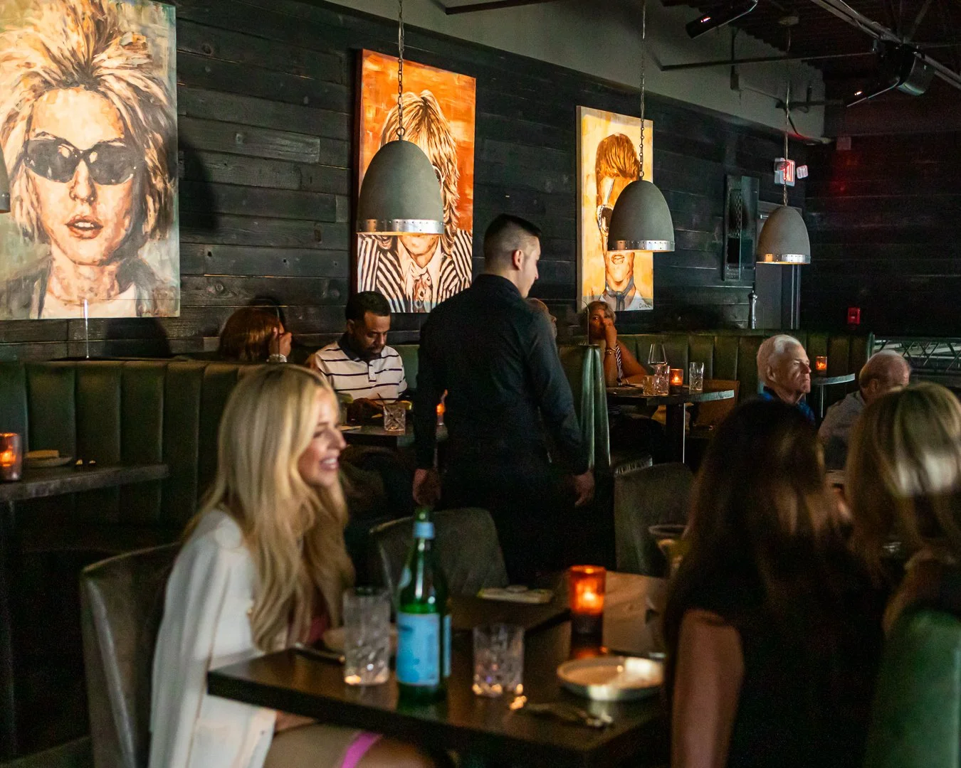 A dimly lit restaurant with black wooden walls decorated with colorful portraits of women. Several patrons sit at tables enjoying food and drinks, and a waiter is serving customers.