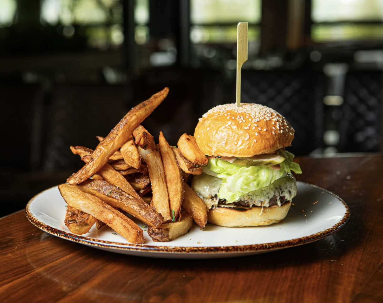 Plate with a cheeseburger topped with sesame seed bun, lettuce, cheese, and a beef patty, served with a side of French fries on a wooden table.