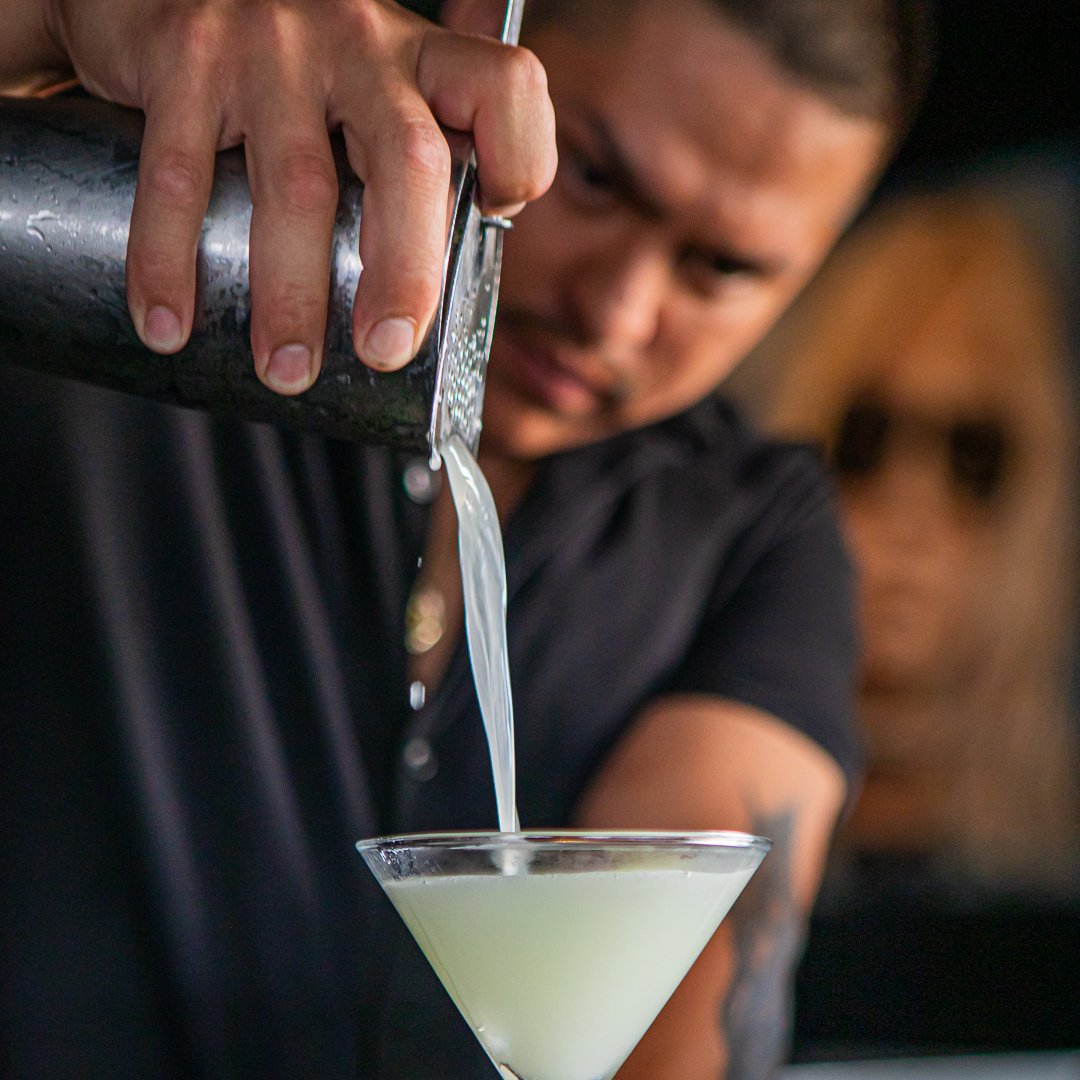 A bartender pours a pale yellow cocktail into a martini glass.