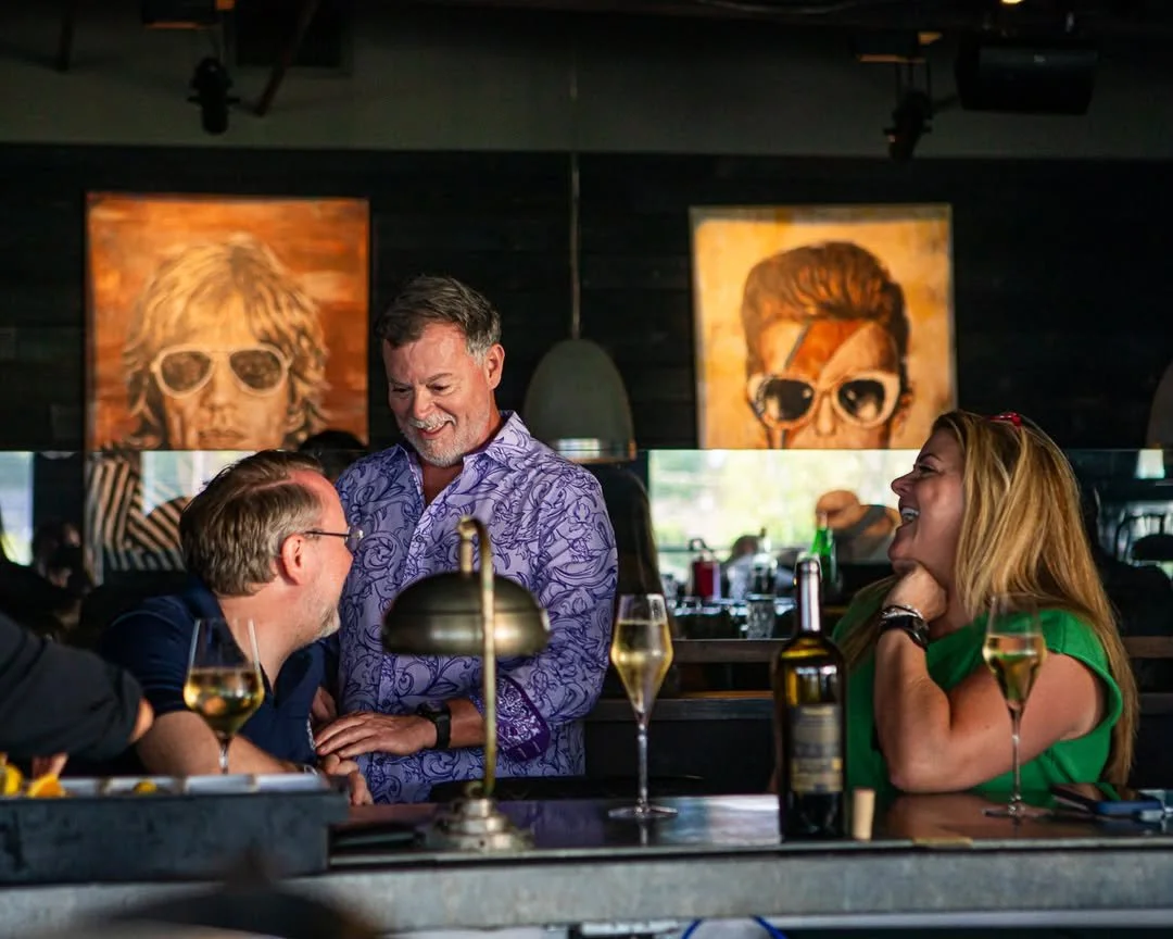 Group of three adults having a lively conversation and laughing at a bar or restaurant with drinks on the counter, black walls, and colorful portraits hanging in the background.