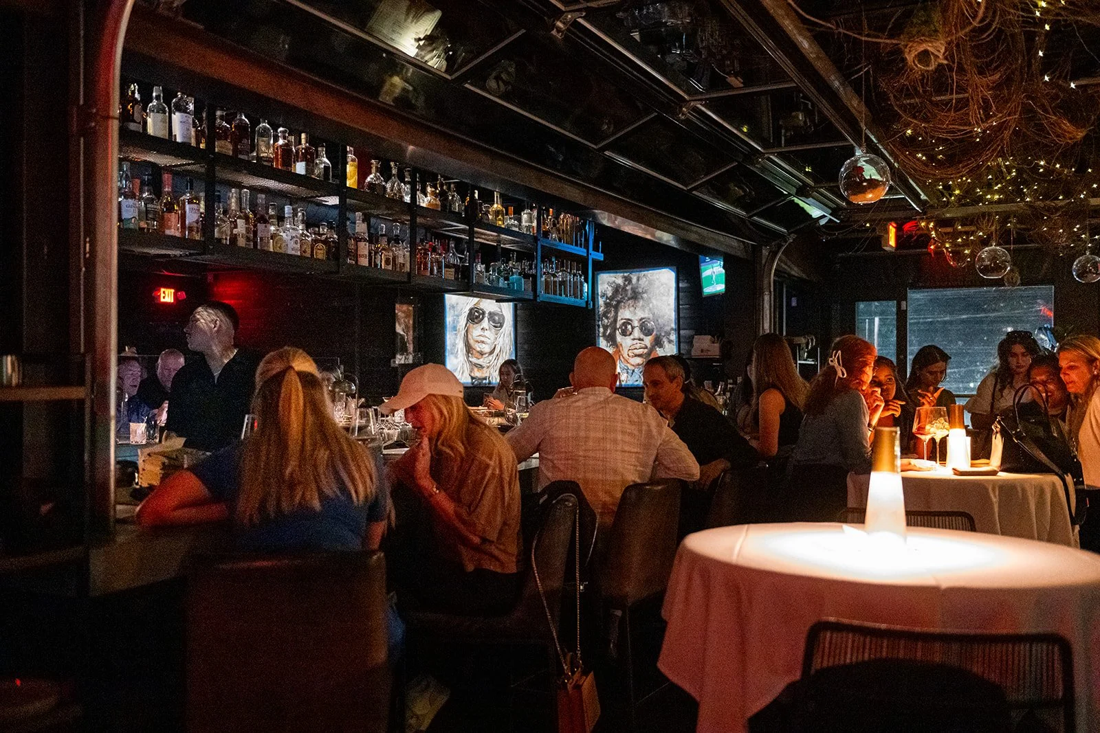A dimly lit bar and restaurant filled with patrons sitting at tables and a bar, with artwork of two people with sunglasses on the wall, and hanging decorative lights.
