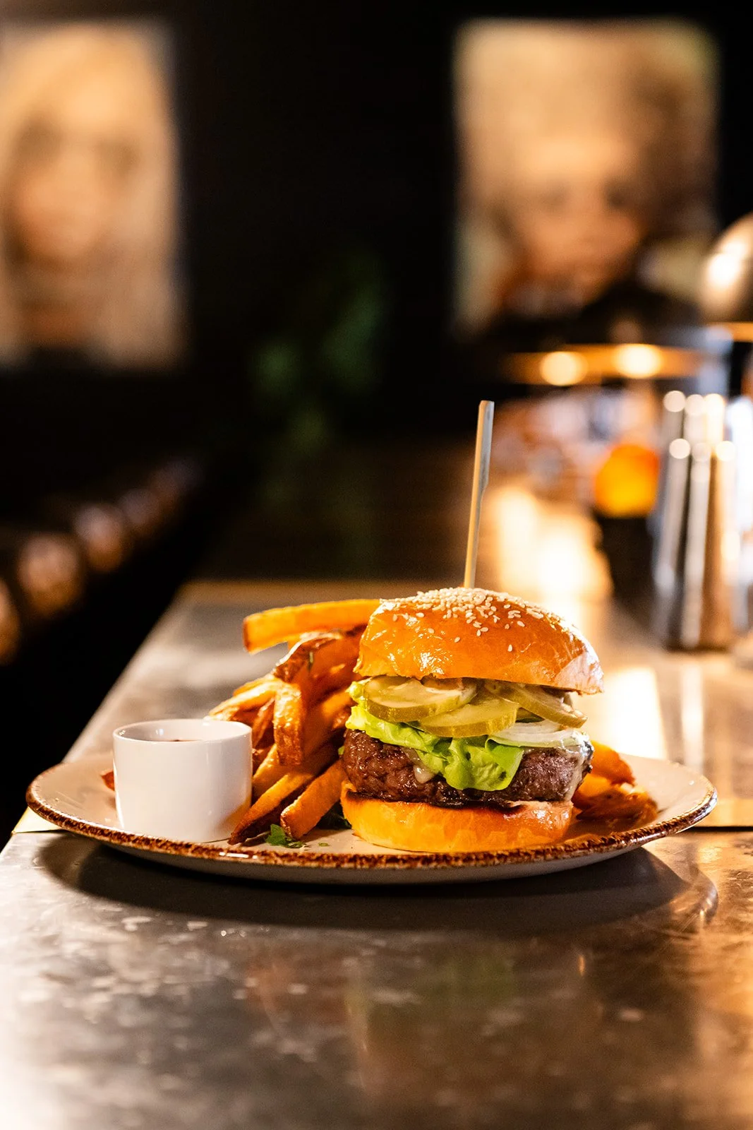 A plate with a cheeseburger, lettuce, pickles, tomato, and a skewer, served with fries and a dipping sauce in a restaurant setting.