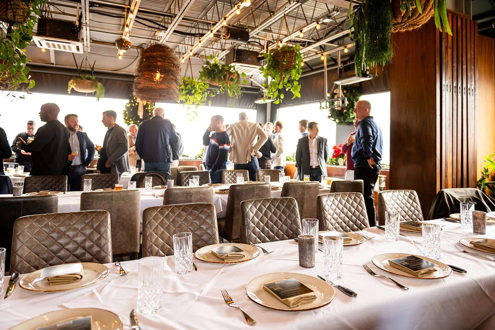 Elegant restaurant or event space with decorated tables set for dining, including gold chargers, glasses, and utensils. People in formal and casual attire are standing and socializing near windows with natural light, greenery, and decorative hanging 