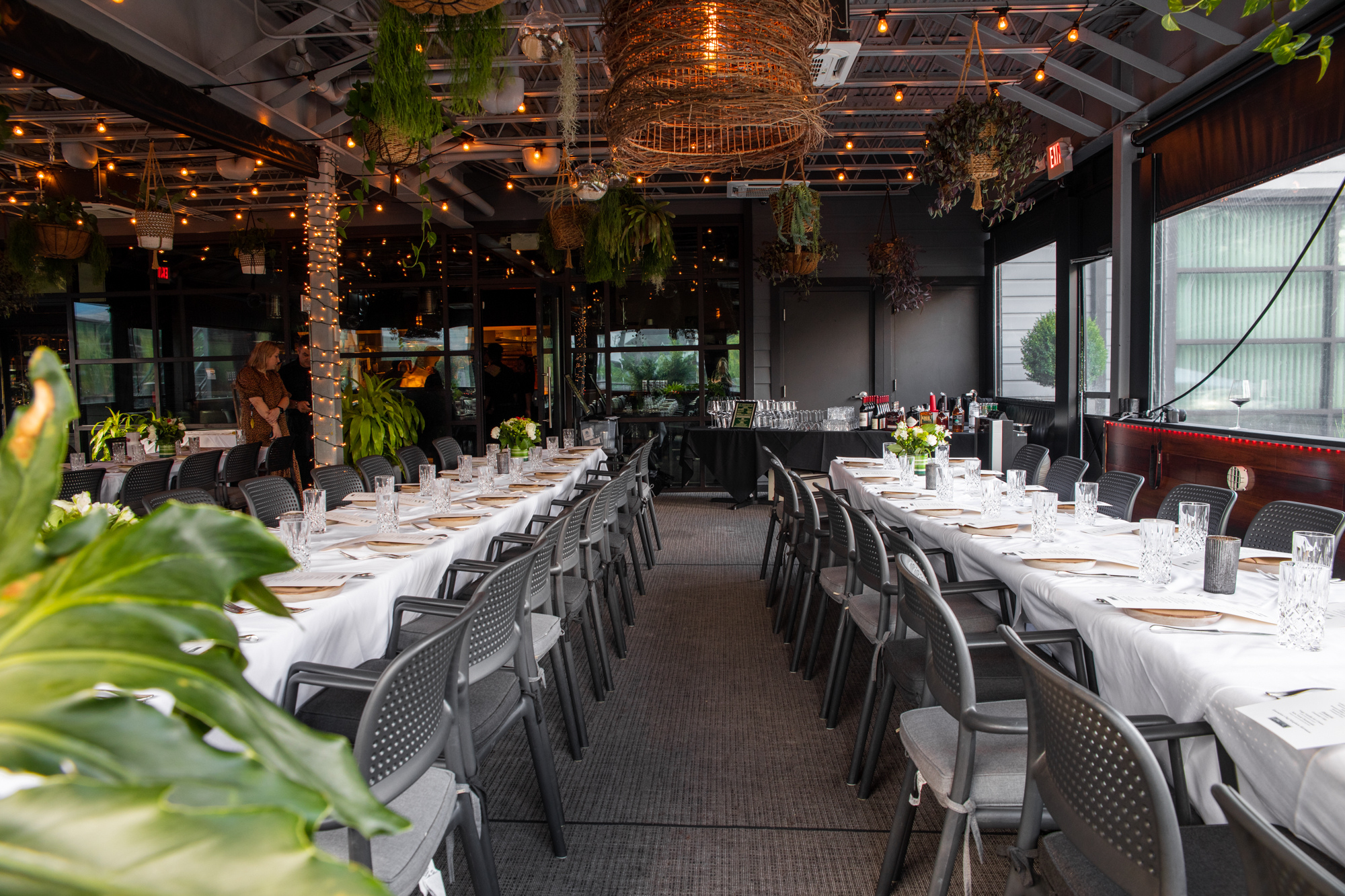 A decorated event venue with long tables covered in white tablecloths, set with glasses, plates, and napkins. There are hanging plants, string lights, and a bar area at the back. The room has large windows and black walls.
