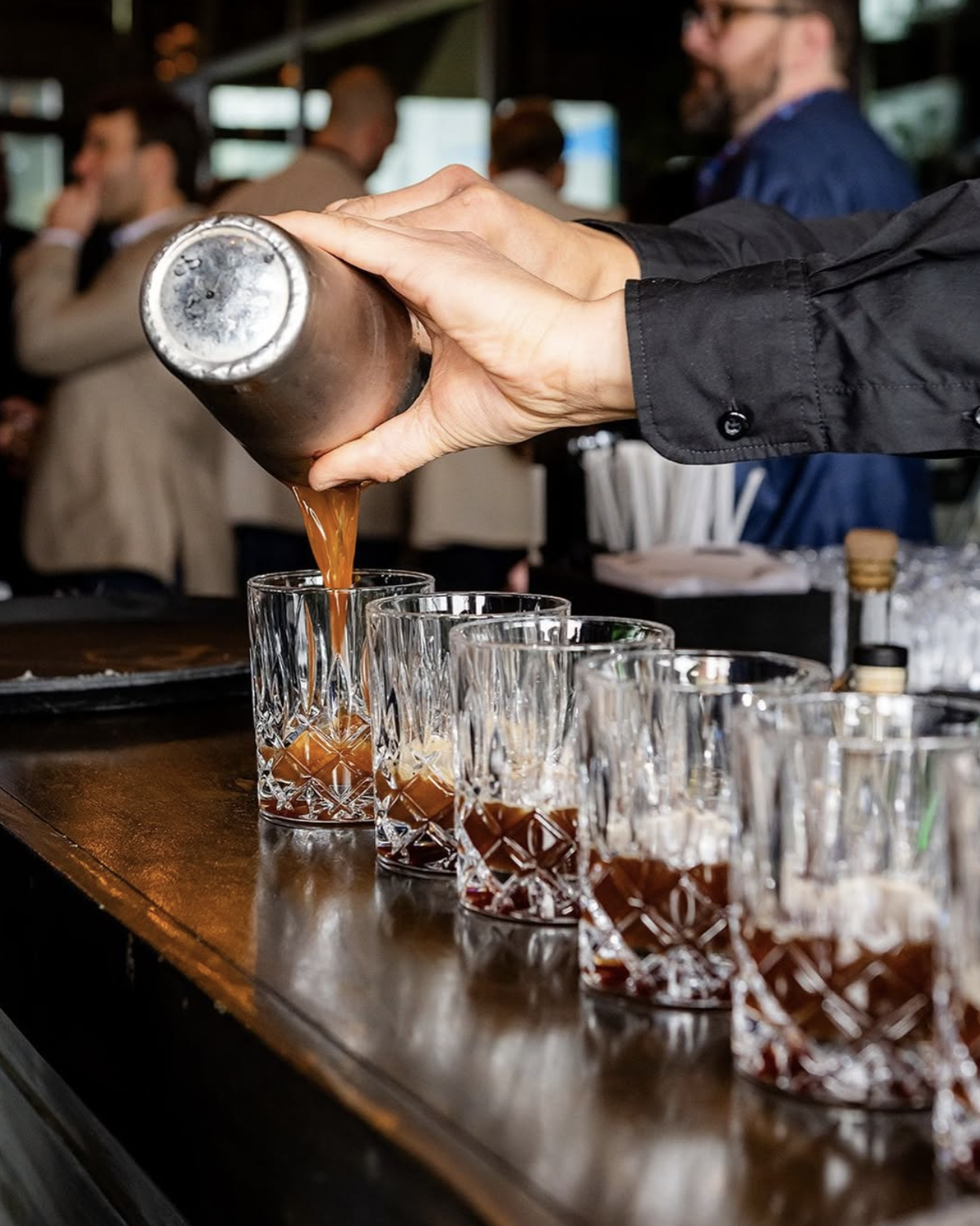 Person pouring a drink into a row of glasses at a bar or event, with a blurred crowd in the background.