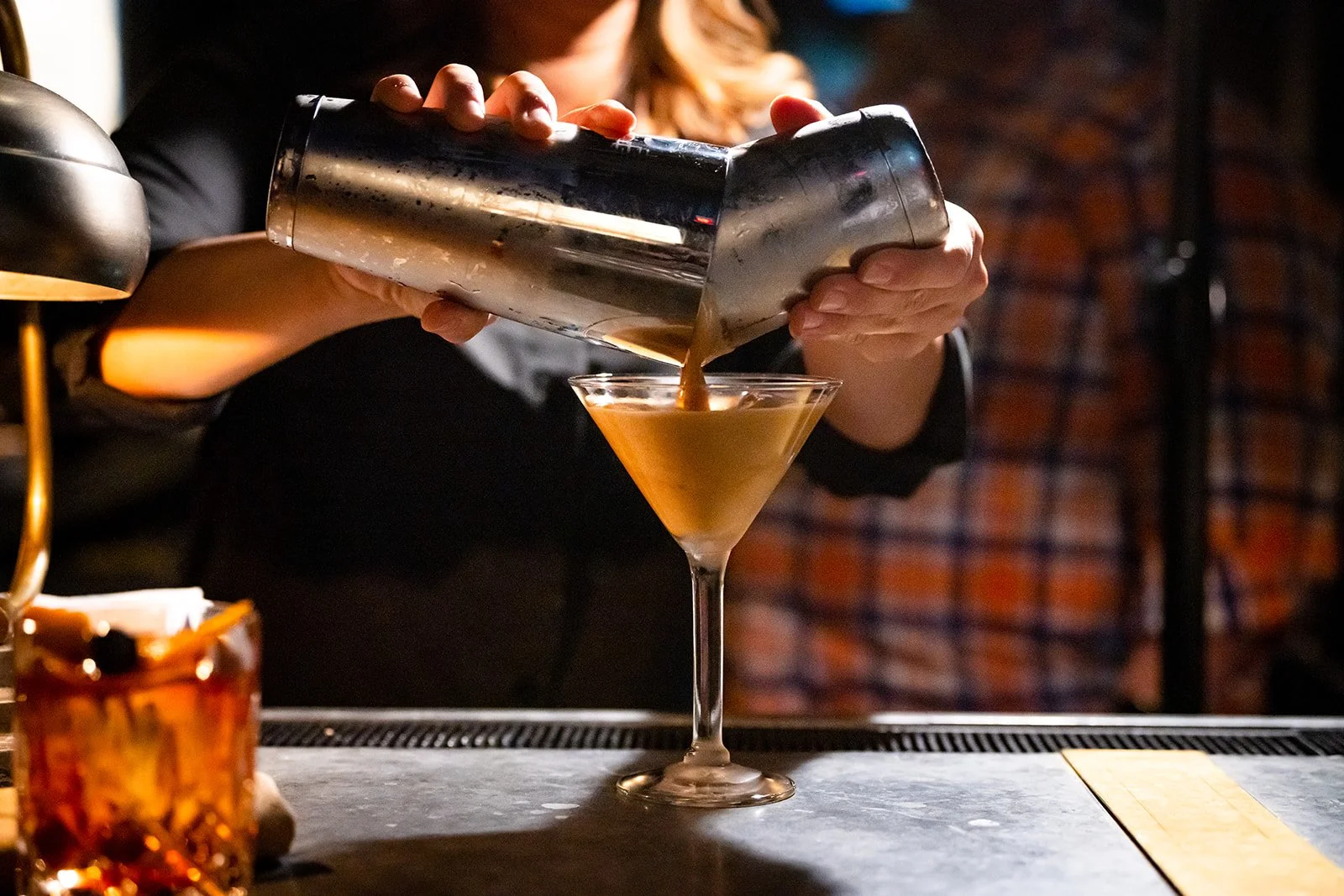 Person pouring brown cocktail from a shaker into a martini glass at a bar.
