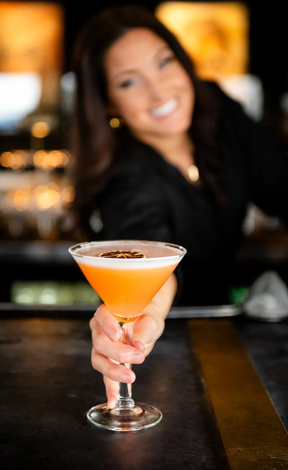 A woman smiling and holding out a pink cocktail garnished with a dried citrus slice in a cocktail glass in a dimly lit bar.