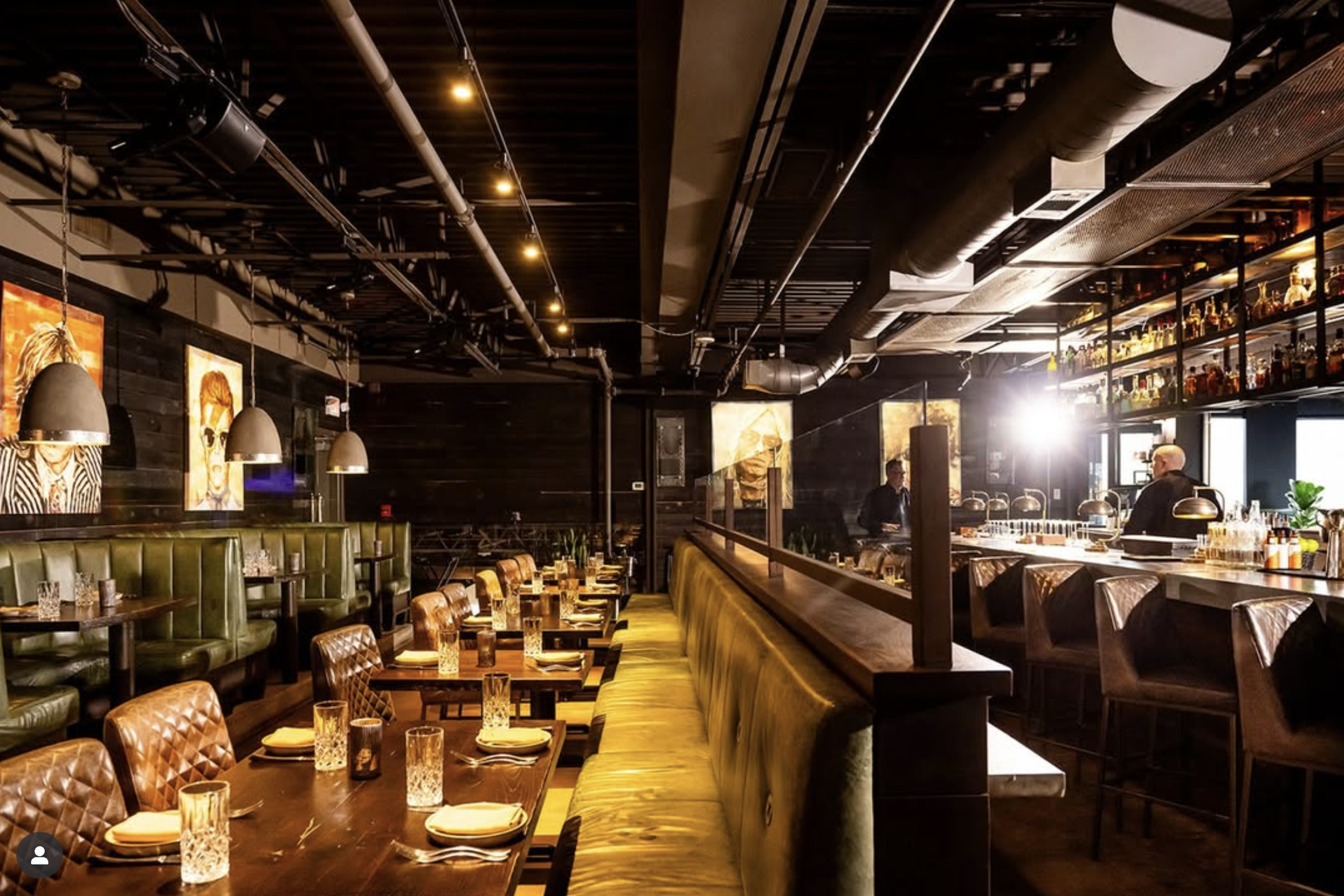 Interior of a stylish restaurant with wooden tables set with glassware and napkins, green leather and quilted brown chairs, dark walls with portraits, and bar area with bottles, illuminated by pendant and ceiling lights.