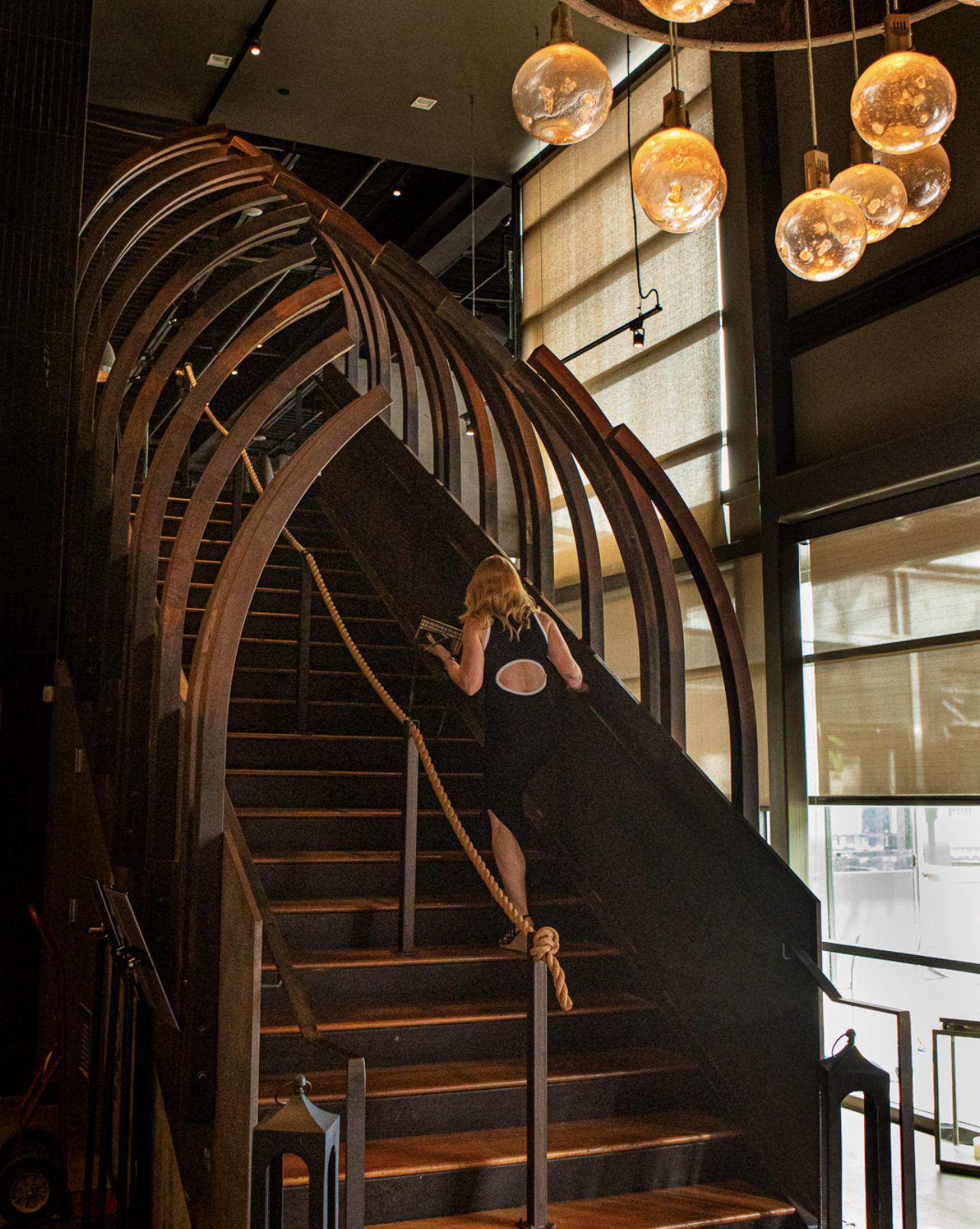 A woman in a black dress with a large cutout on the back climbs a dark wooden staircase in a modern interior with large windows and spherical hanging light fixtures.