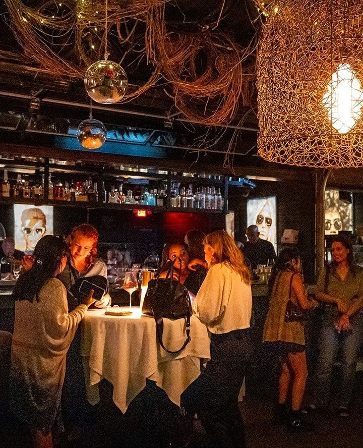 People socializing around a table at a dimly lit bar or restaurant with festive decorations, including hanging mirrors and woven light fixtures.
