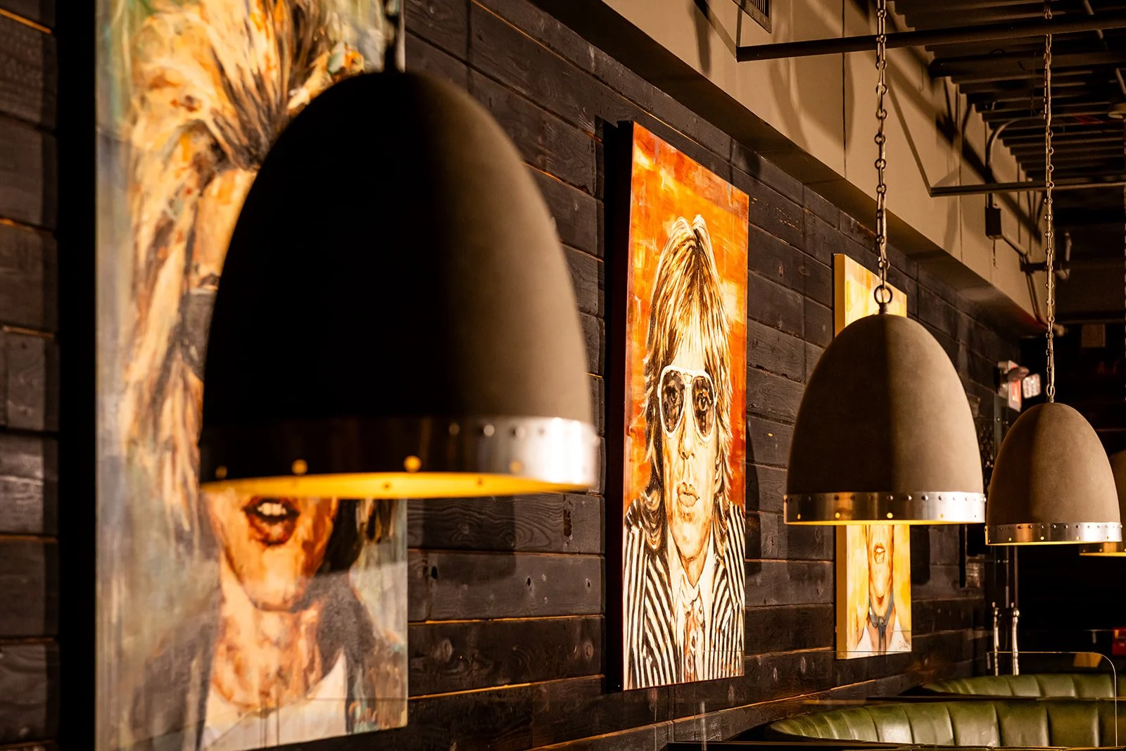 Interior of a restaurant or cafe with black wall paneling, featuring colorful portraits of women with glasses, illuminated by large black pendant lights.