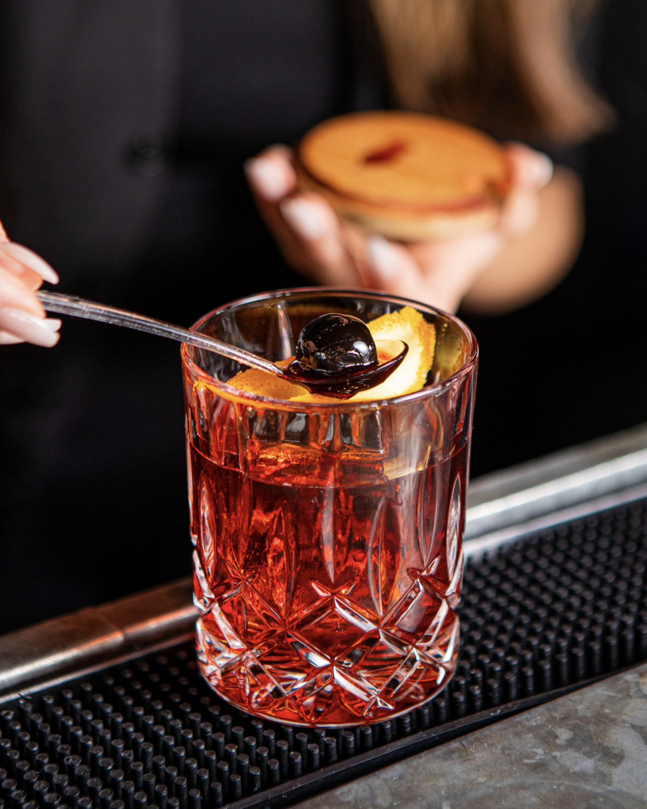 A bartender adds a cherry to a red cocktail with orange slices in a cut-glass tumbler.