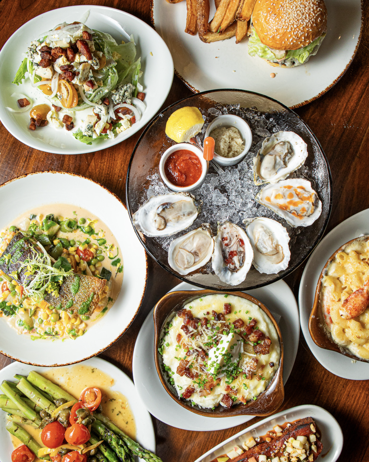 Assorted dishes including a seafood platter with oysters, lemon, cocktail sauce, and horseradish; a grilled fish with vegetables; a baked pasta casserole; a salad with bacon and cheese; a plate of asparagus and cherry tomatoes; a cheeseburger with fr