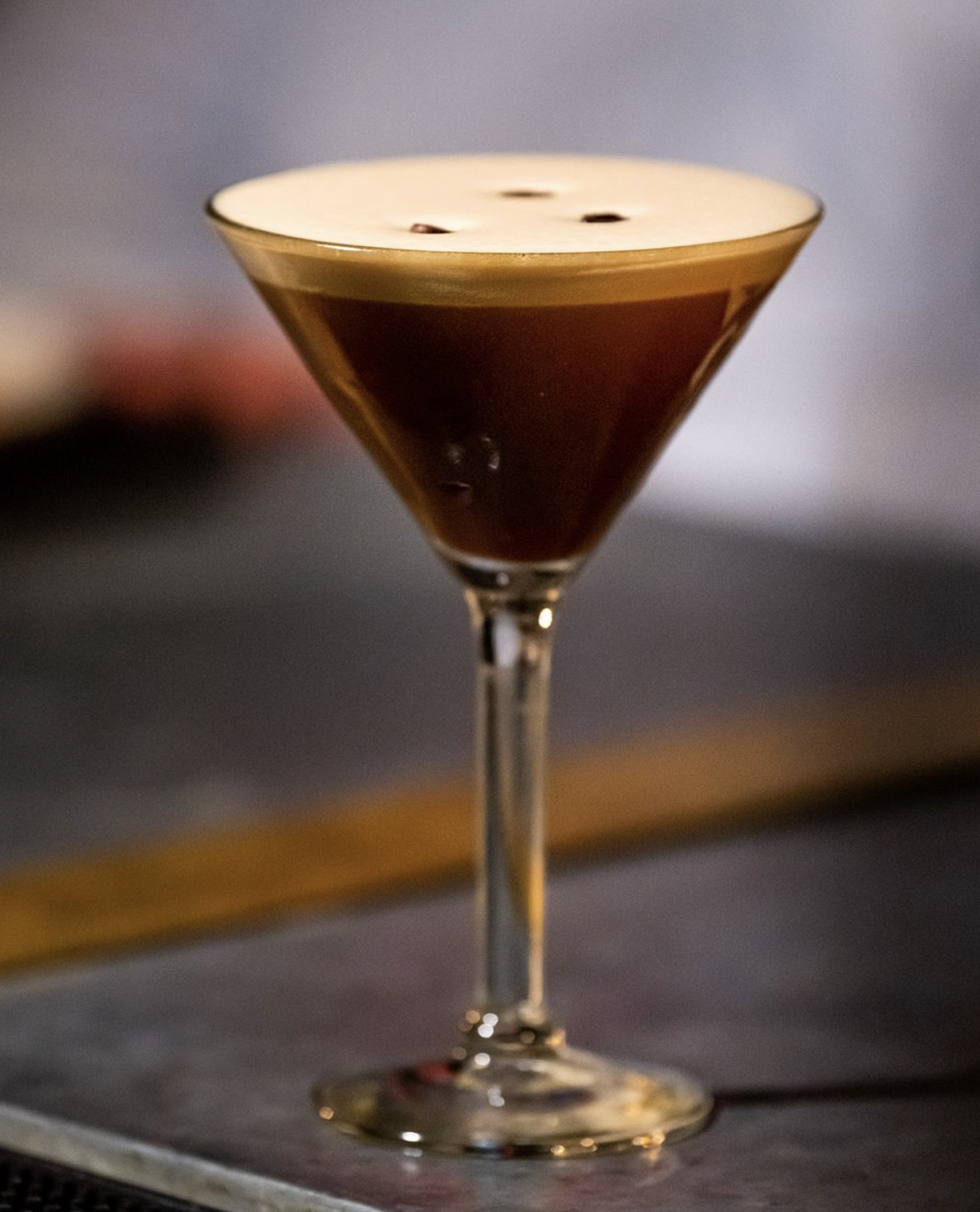 Close-up of an espresso martini cocktail in a tall, slender martini glass with a creamy froth and three coffee beans on top.