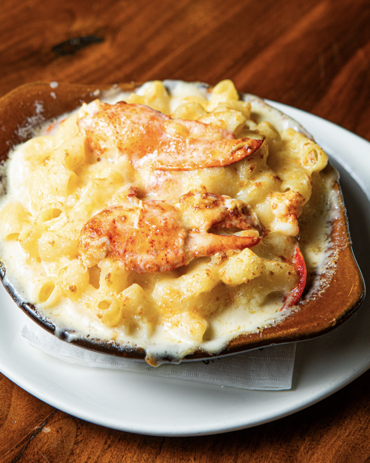 Baked pasta with cheese, lobster, and red bell peppers served in a ceramic dish on a white plate.