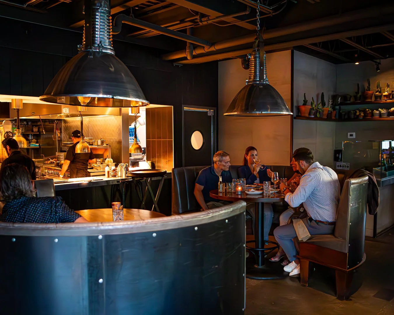 A cozy restaurant with dim lighting, showing customers dining at a booth, restaurant staff working behind the counter, and potted plants on shelves in the background.
