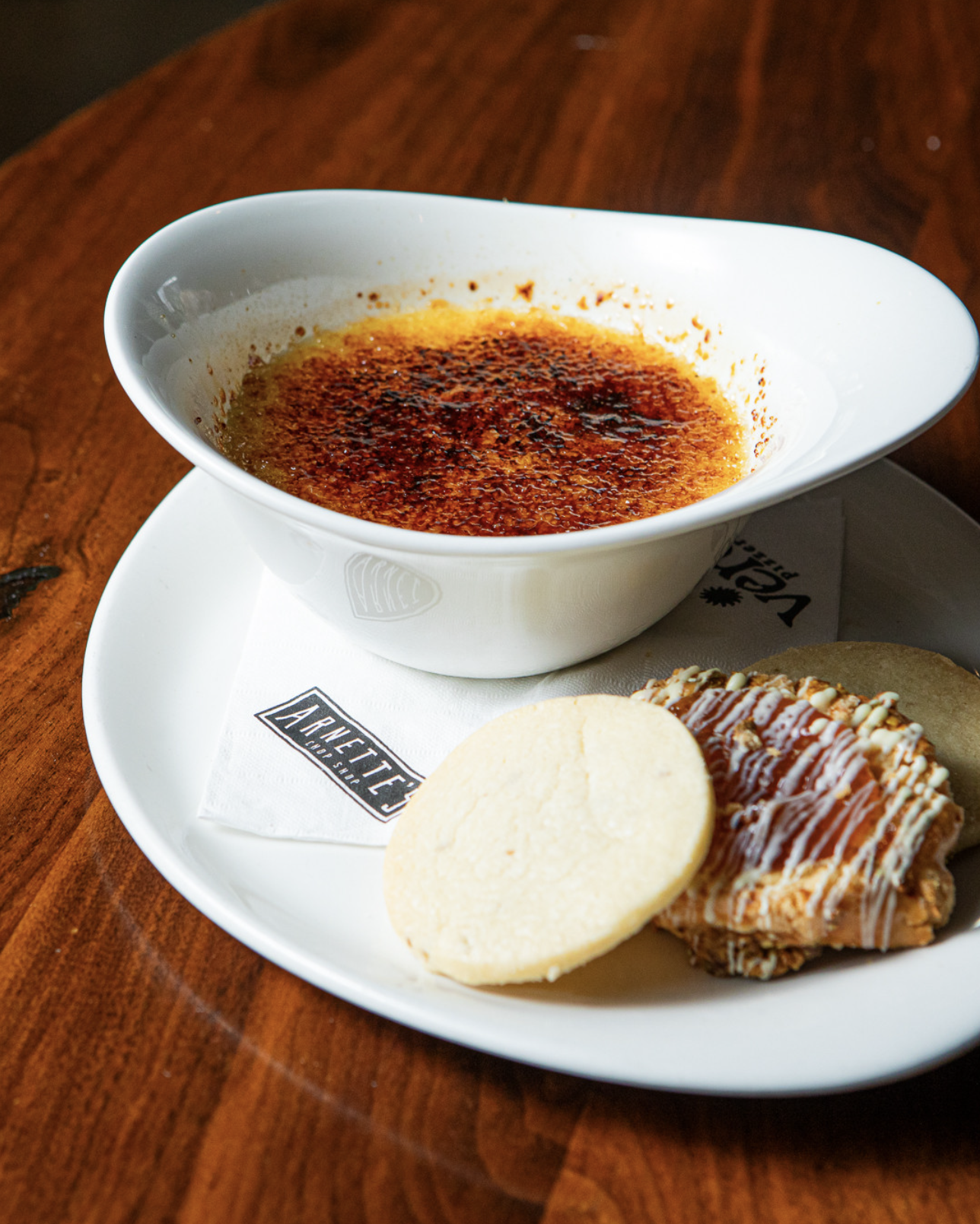 Crème brûlée dessert with a caramelized sugar crust on top, served in a white bowl on a white plate, accompanied by a round shortbread cookie and an almond-covered pastry drizzle with white chocolate.
