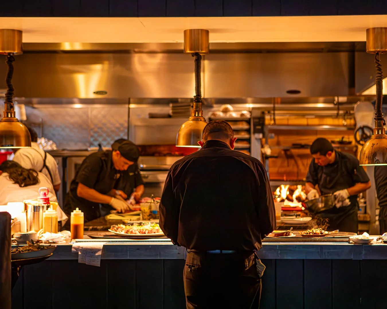Chef working at a busy restaurant kitchen with multiple chefs preparing dishes and a man observing.