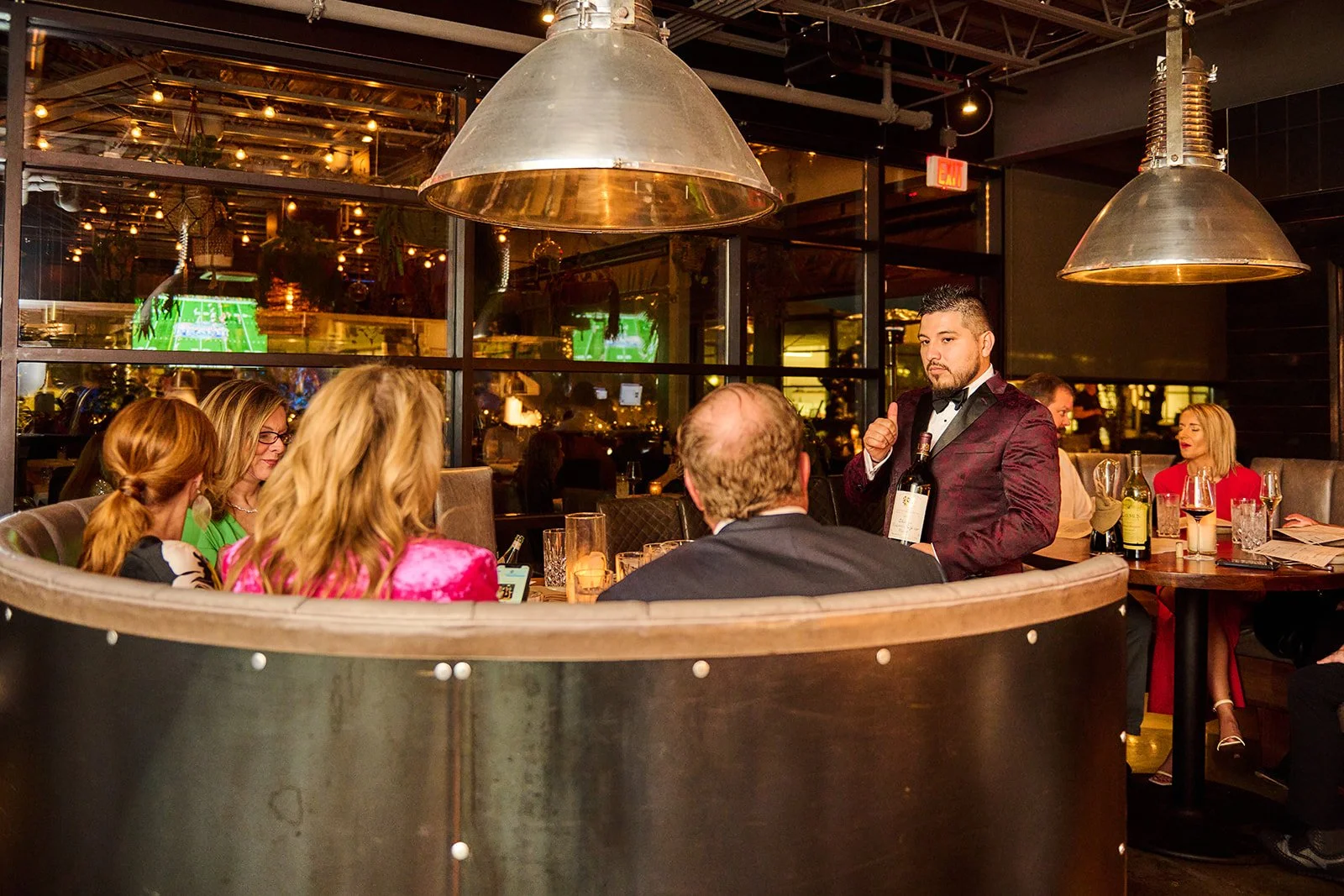 A group of people dining in a restaurant, with a server in a tuxedo serving drinks. The restaurant has warm lighting, large metal pendant lamps, and a modern industrial decor.
