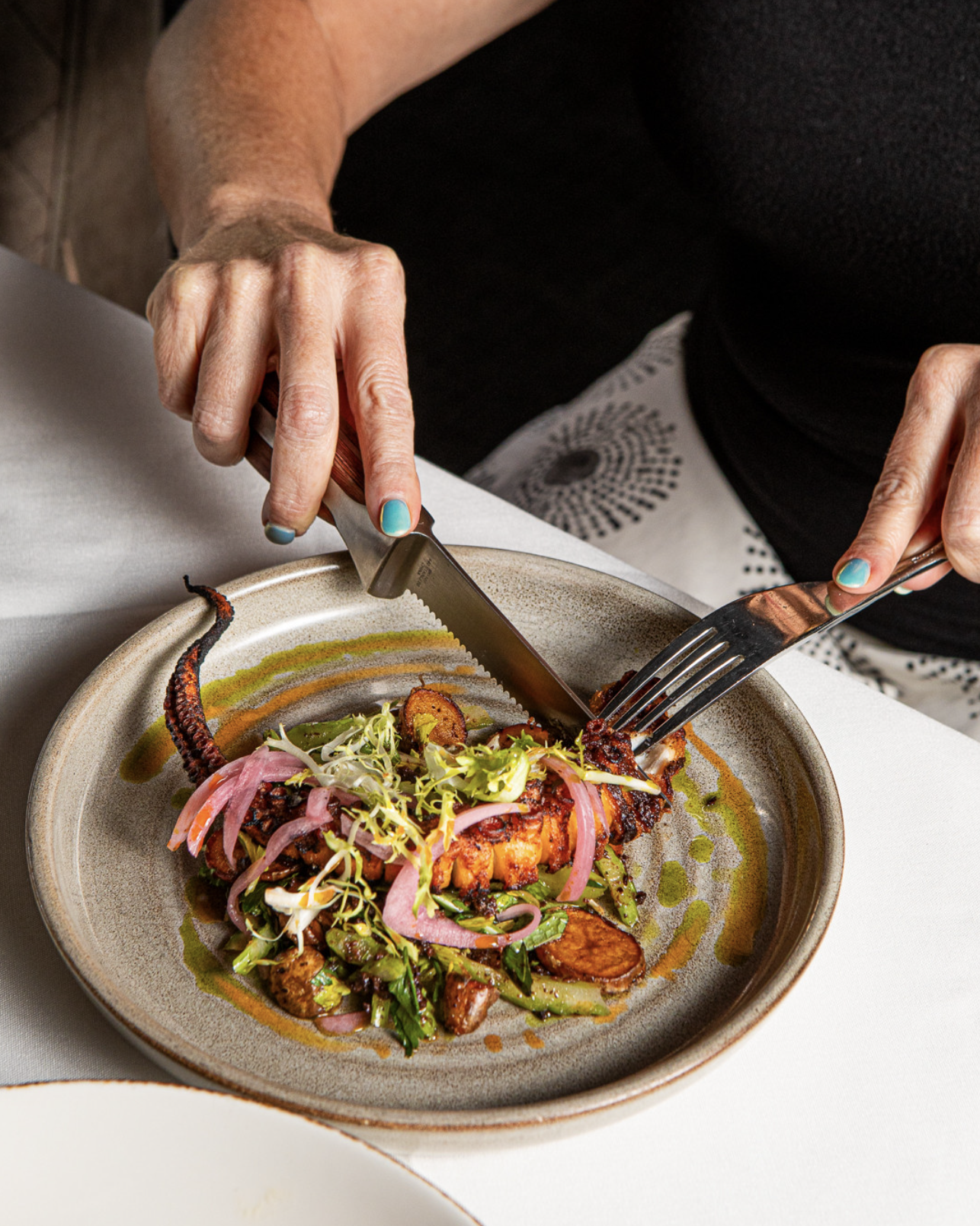 Person cutting into a grilled octopus tentacle on a plate with vegetables and garnishes.