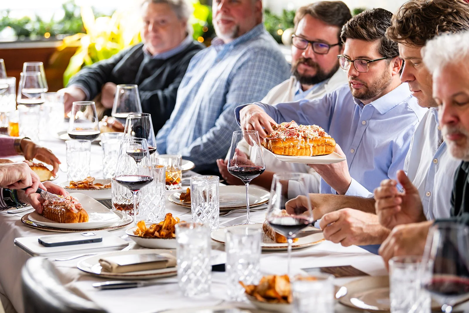 Group of people sitting around a dinner table, sharing a meal, with some holding glasses of red wine and plates of lasagna, in a bright, festive setting.