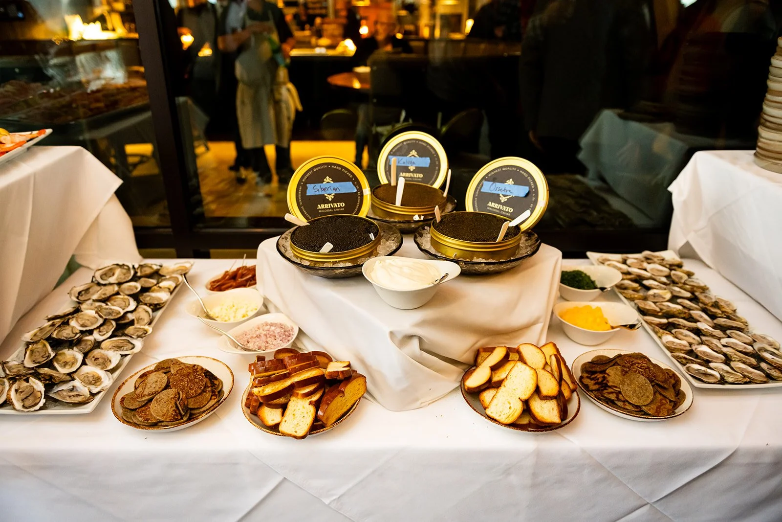 A seafood buffet display with oyster shells, bread slices, and various toppings, set on a white tablecloth. In the background, a glass window reveals a restaurant interior with people dining.