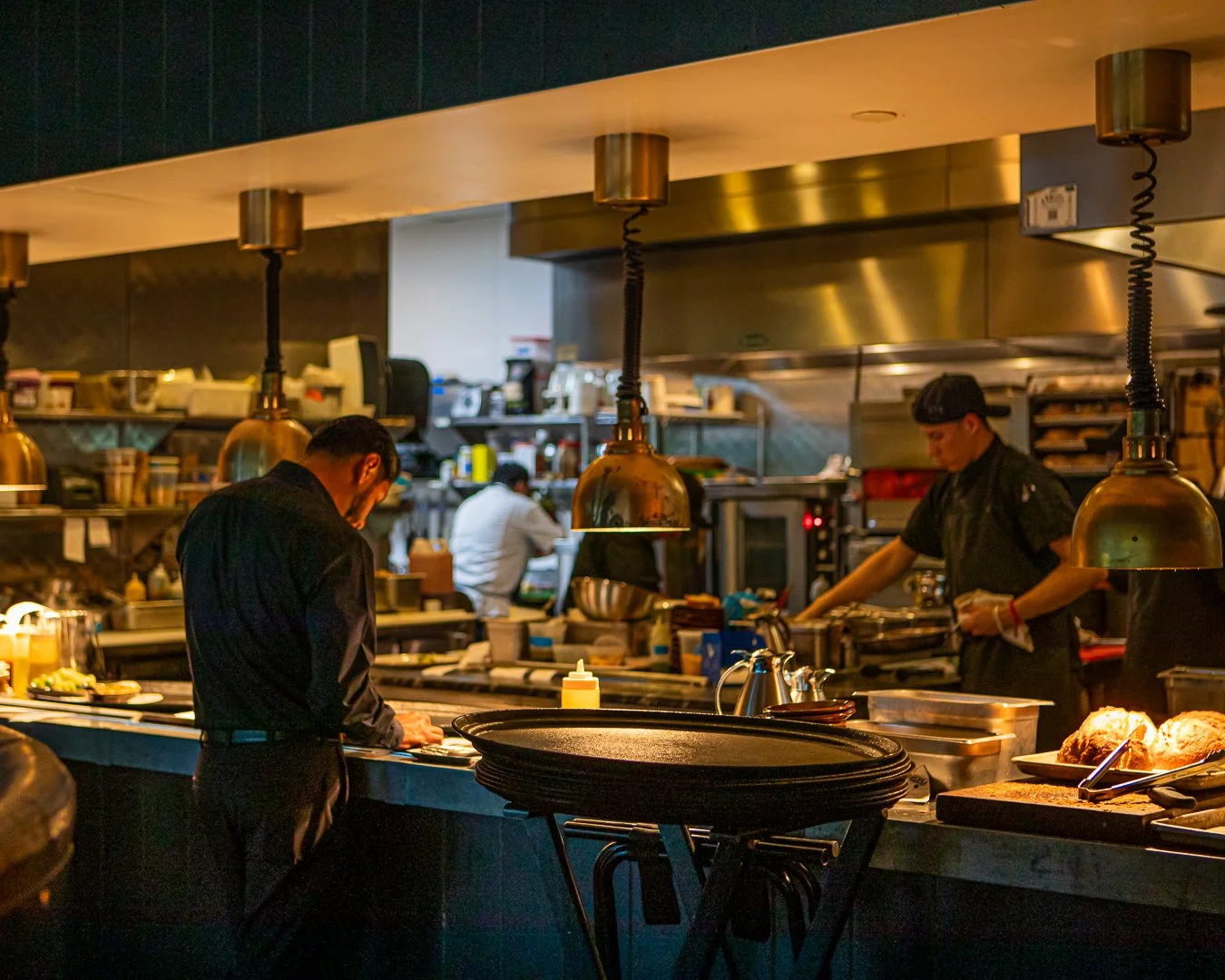 Inside a busy restaurant kitchen with chefs preparing food behind a counter, with hanging lights over the workspace.