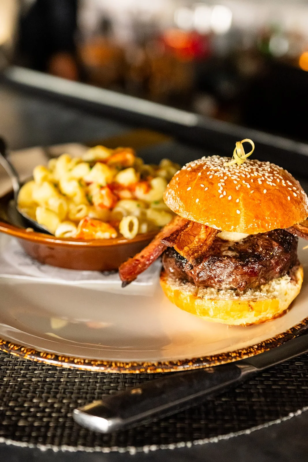 A burger with bacon, beef patty, and bun served with a side of pasta salad on a white plate.