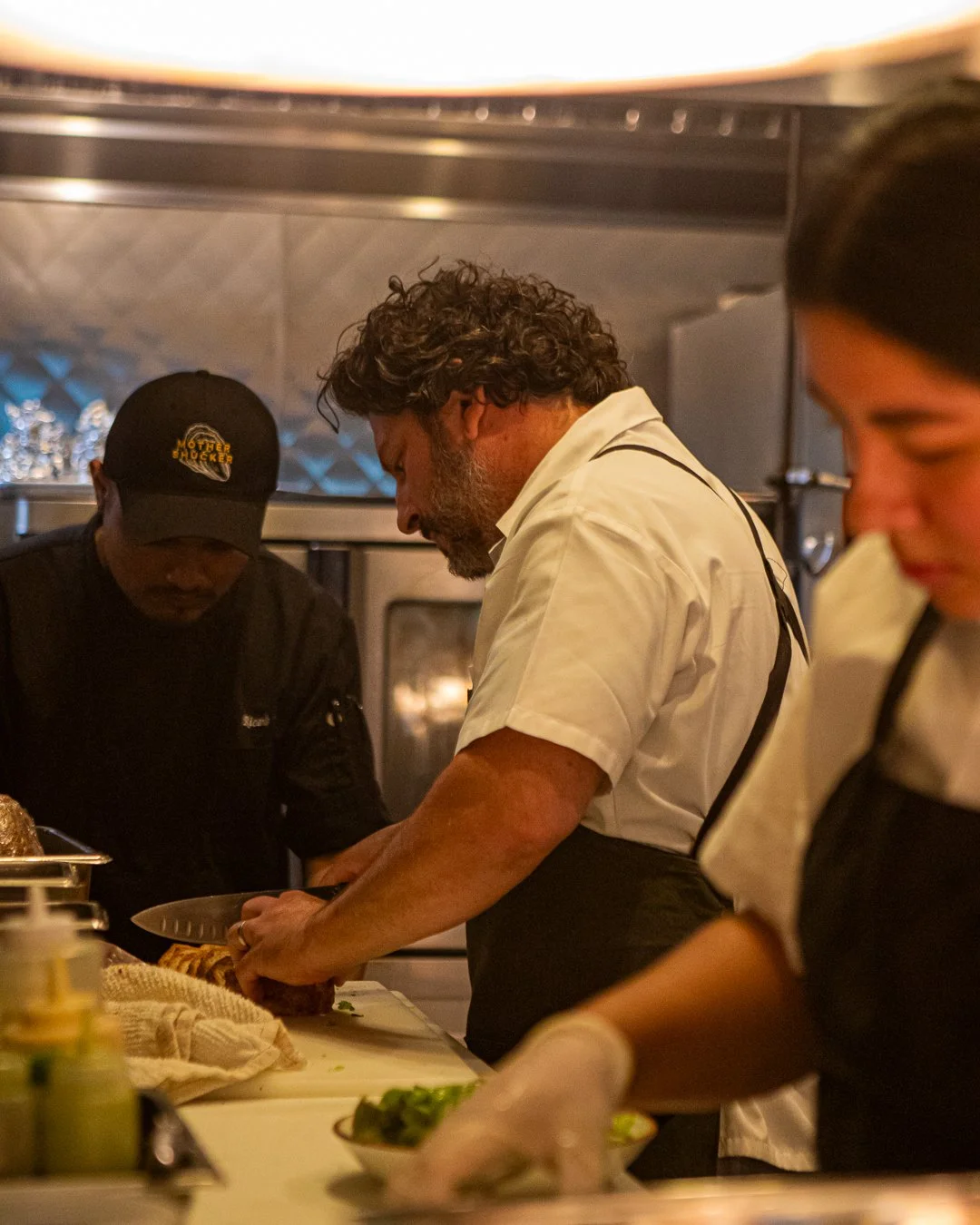 Chef slicing meat in a busy commercial kitchen with two other staff members preparing ingredients nearby.