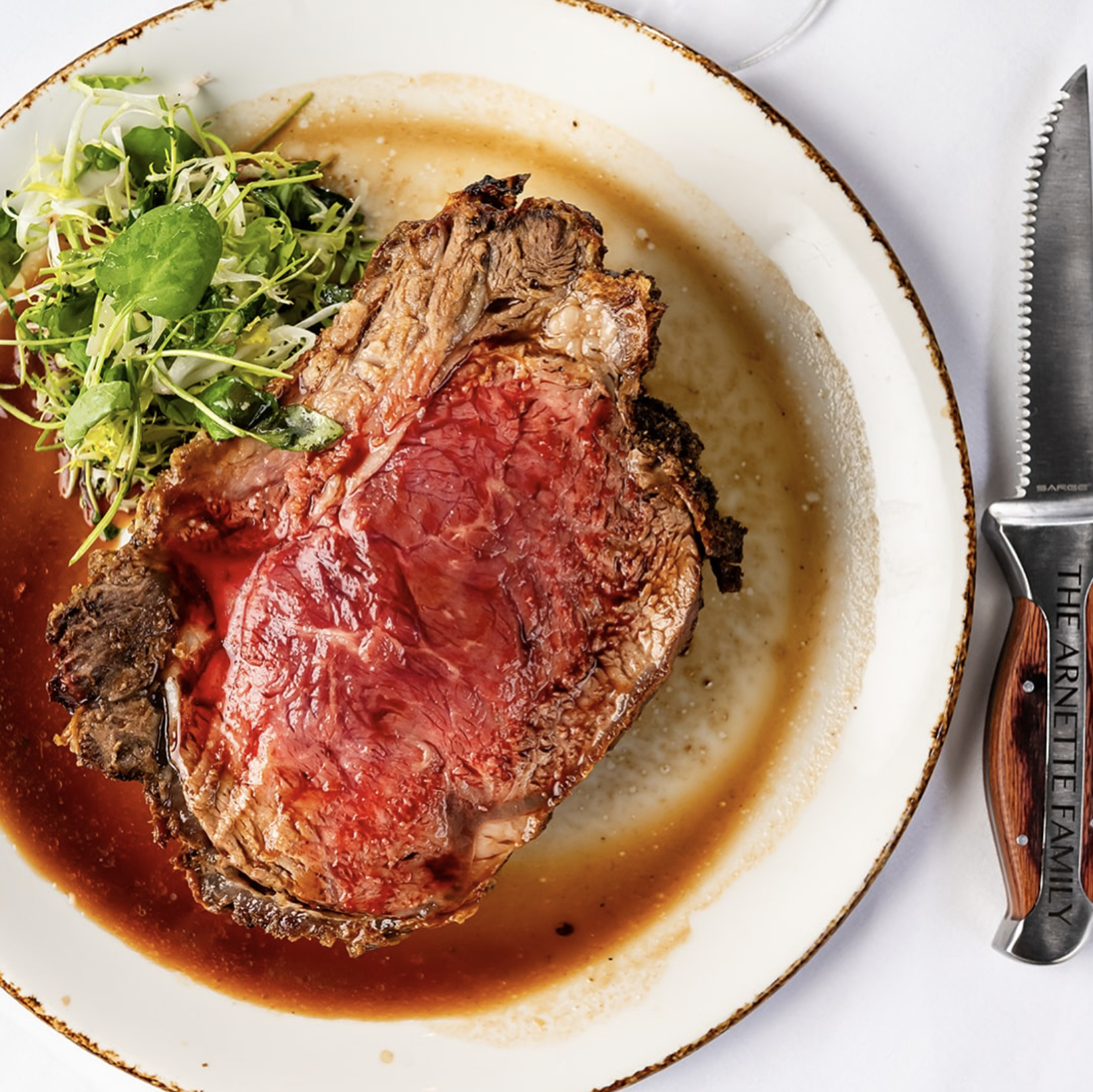 A cooked prime rib roast with a medium-rare interior, set on a white plate with some its juices. There is a side of green salad with arugula and microgreens on the plate. A carving knife with a wooden handle labeled 'The Farnet Te Family' is placed b